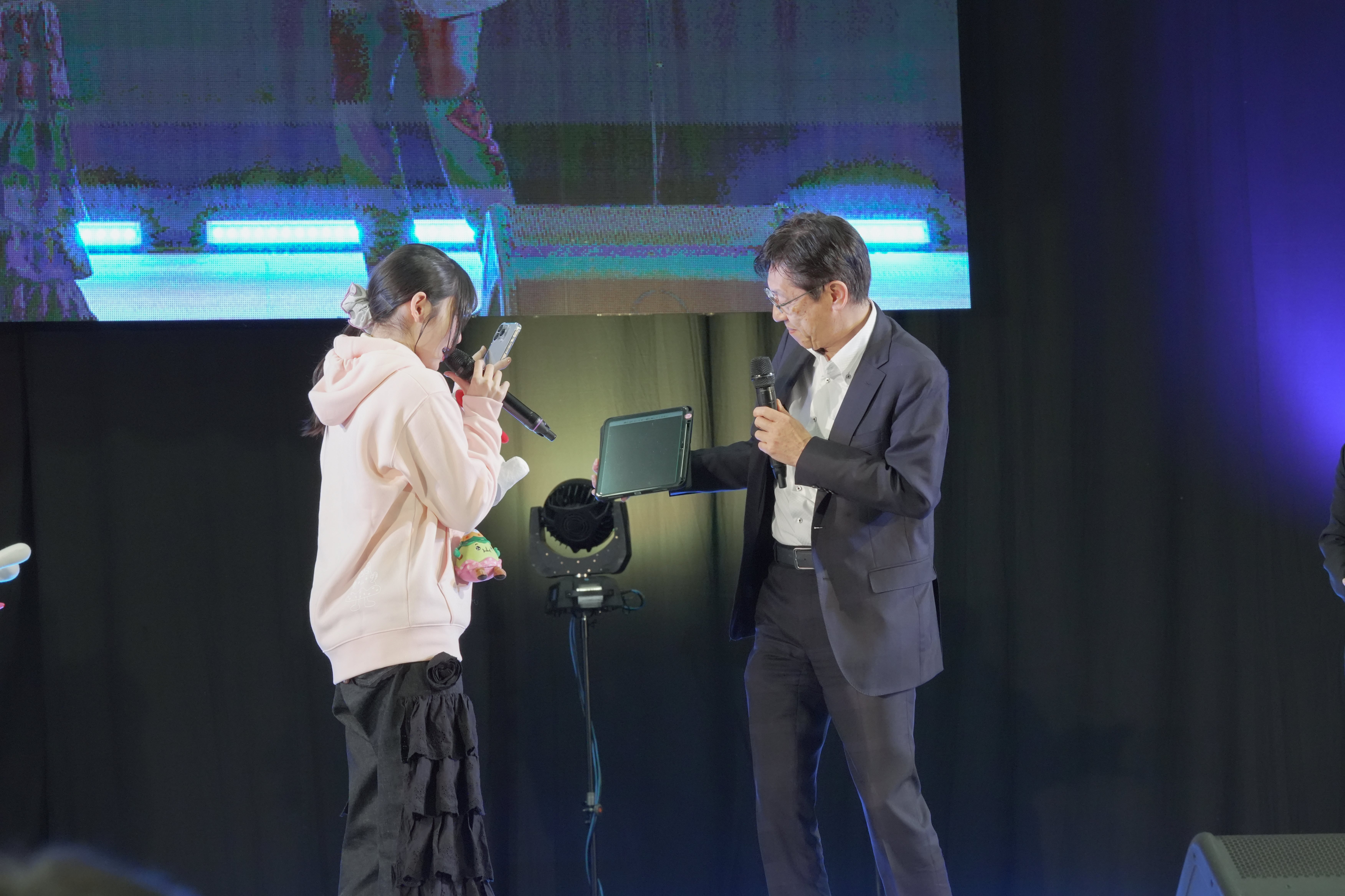 暗転したステージ上で、ピンクのパーカーと黒いフリルスカートを着た若い女性が、マイクとスマートフォンを手に持ち、画面を確認している。向かいに立つ眼鏡をかけたスーツ姿の松本デジタル大臣がマイクを持ちながらタブレット端末を差し出している。背後には青いLEDライトと大型スクリーンがある。