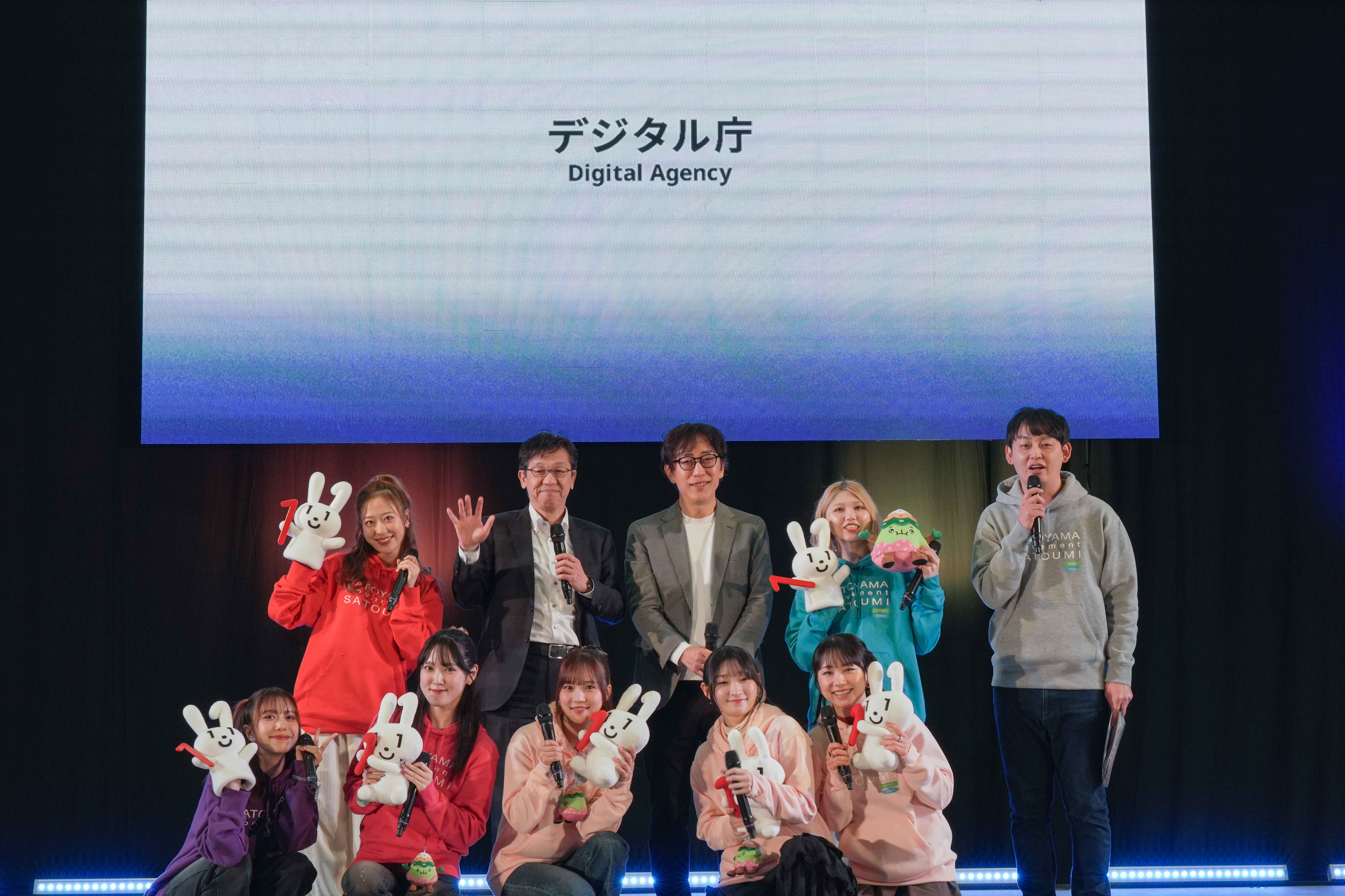 ステージ上で総勢10名程度が並んでいる。背後の大型スクリーンには『デジタル庁 Digital Agency』の文字が表示されている。後列にはスーツ姿の男性2名とマイクを持った男性1名が立ち、前列には左から紫と赤のパーカーを着た女性がそれぞれ1名、ピンクのパーカーを着た女性3名の計5名が座っている。多くの出演者が白いマイナちゃんのぬいぐるみを手に持ち、笑顔でカメラに向かっている。ステージ前面には青いLEDライトが並んでいる。