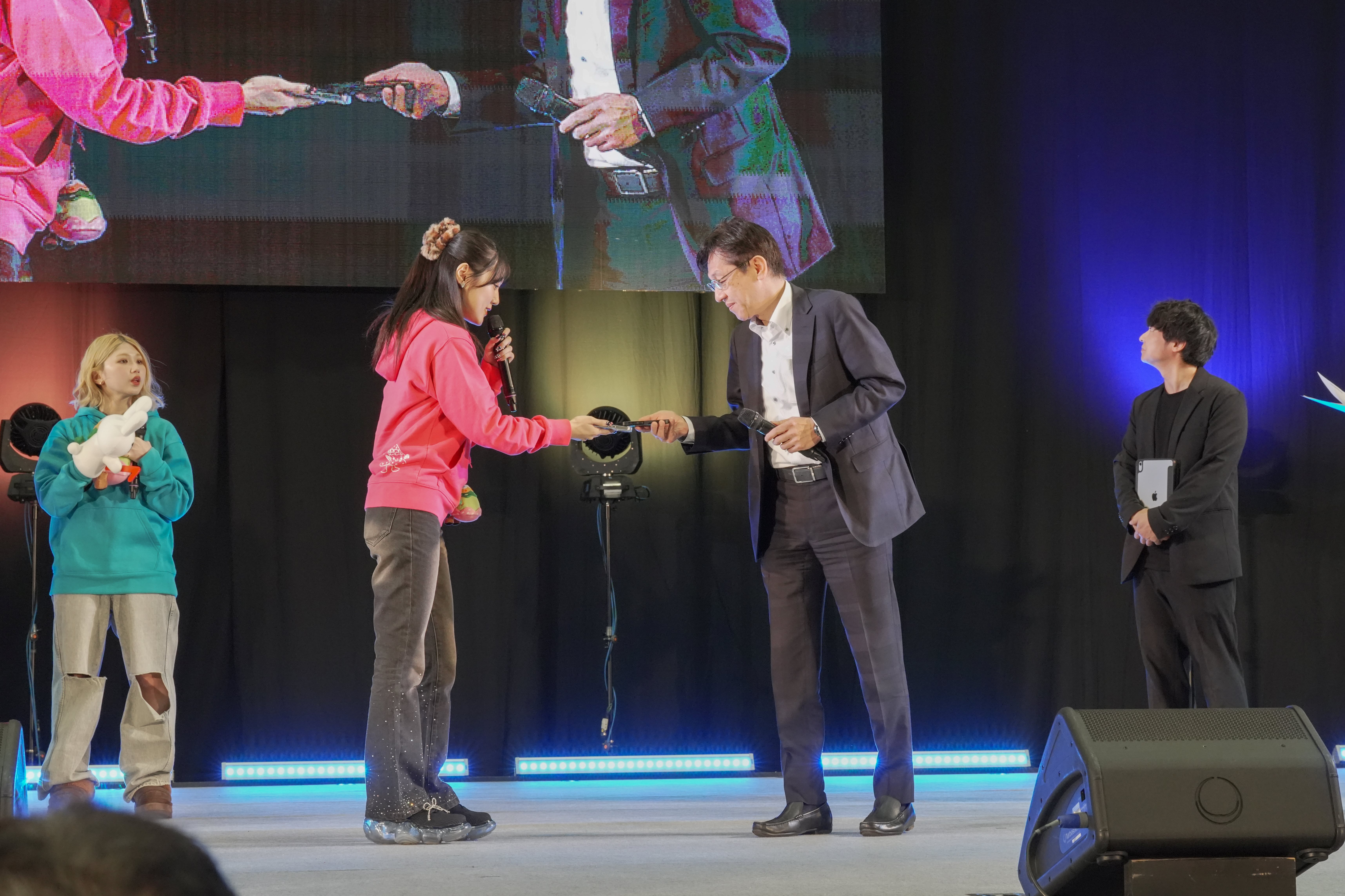 ステージ上で、ピンクのパーカーを着てマイクを持った女性が、スーツ姿の眼鏡をかけた松本デジタル大臣に向かって前かがみになり、スマートフォンをかざしている。松本デジタル大臣も前かがみになってスマートフォンをかざしている。背後の大型スクリーンには二人がスマートフォンをお互いにかざす映像が映し出されている。左側には水色のパーカーを着た女性が白いマイナちゃんのぬいぐるみを抱えて立っており、右側には黒い服装の男性が白いタブレットを持って立っている。ステージ前面には青いLEDライトが並んでいる。