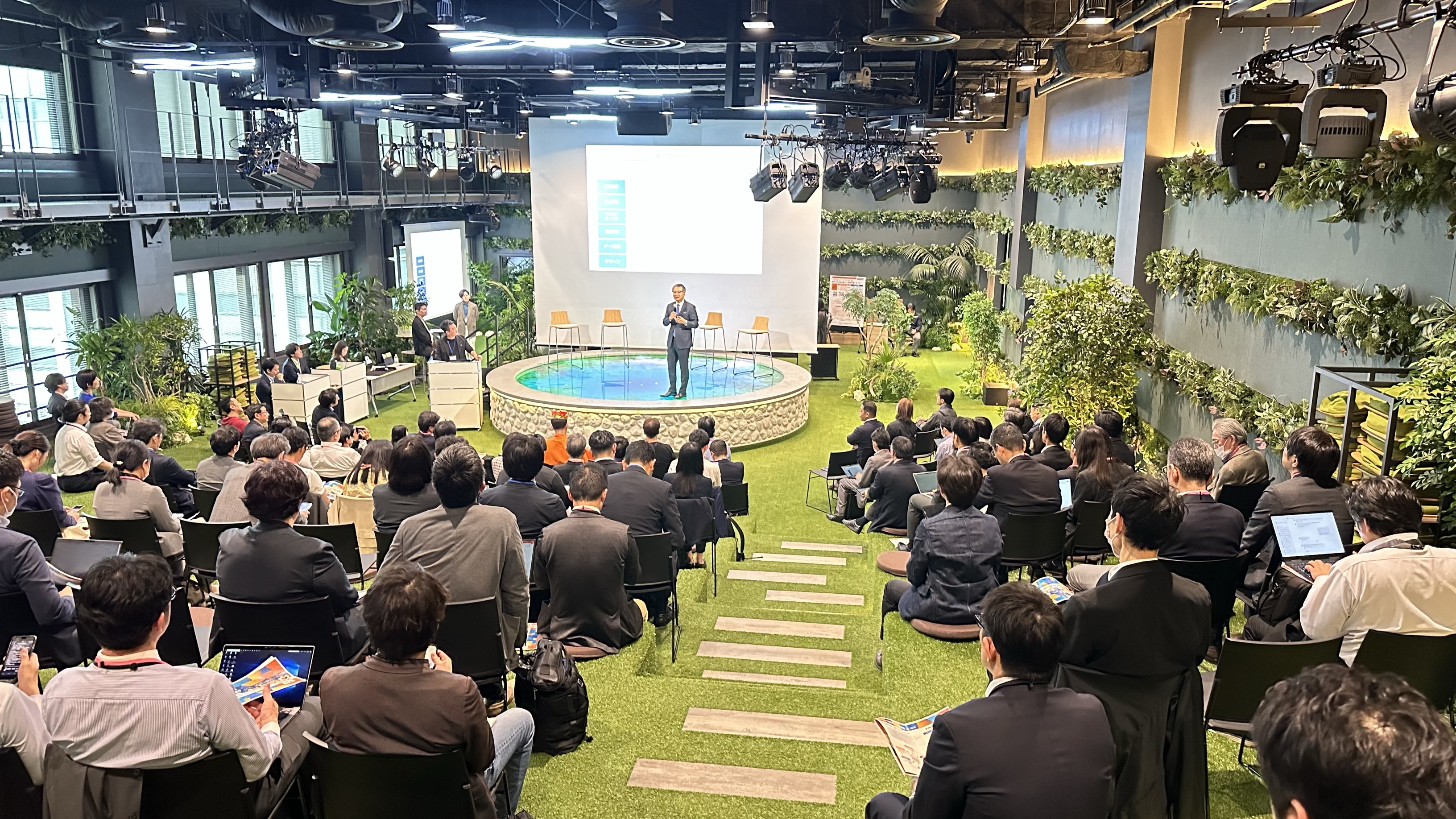A scene of the whole hall. The presenter is standing and talking to the audience. Behind the presenter, there is a projector that projects materials. The floor is stepped and covered with grass. Many spectators participate and sit on the steps and chairs made by the grass to listen to the story.