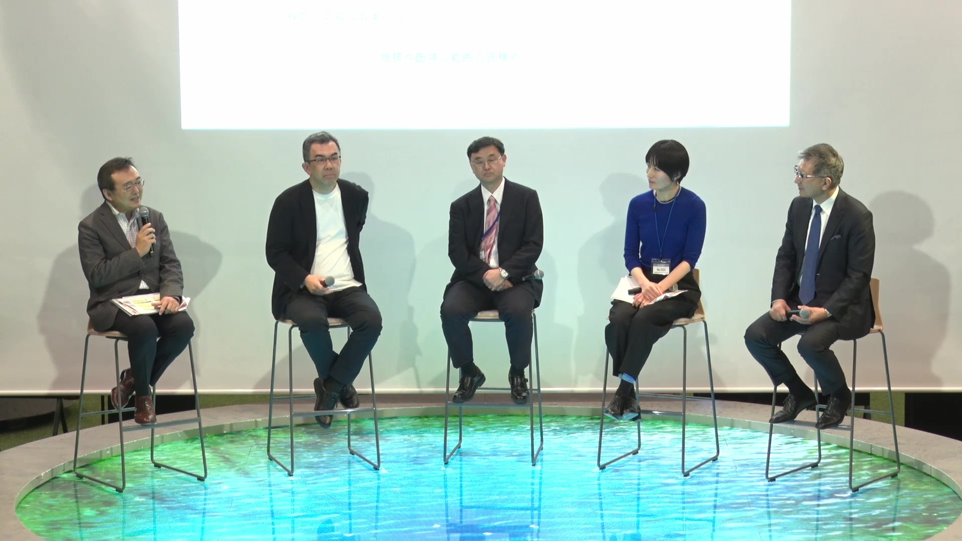 Five people are seated side by side facing the audience seats. The moderator, Director-General Digital Agency Murakami, is holding a microphone and speaking. Behind the panelists, there is a projector that projects the material.