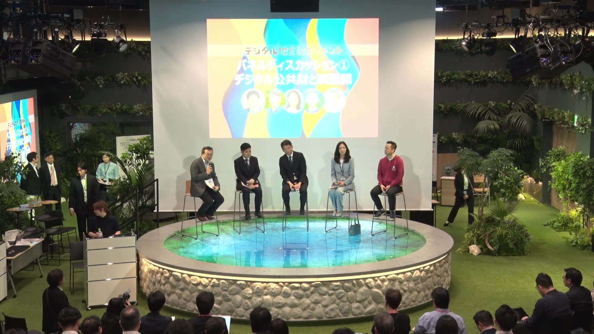 Five people are seated side by side facing the audience seats, and Director-General MURAKAMI is speaking through a microphone. There is a projector behind the panelists to project materials. The venue is decorated with grass on the floor and various green plants on the walls and floor.
