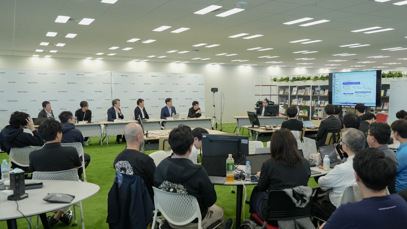 デジタル庁の平将明デジタル大臣ら6名がコメンテータとなり、イベントで開発されたAIのプロトタイプを講評している。会場の前方にコメンテータが並んで着席し、参加者が講評に耳を傾けている。
