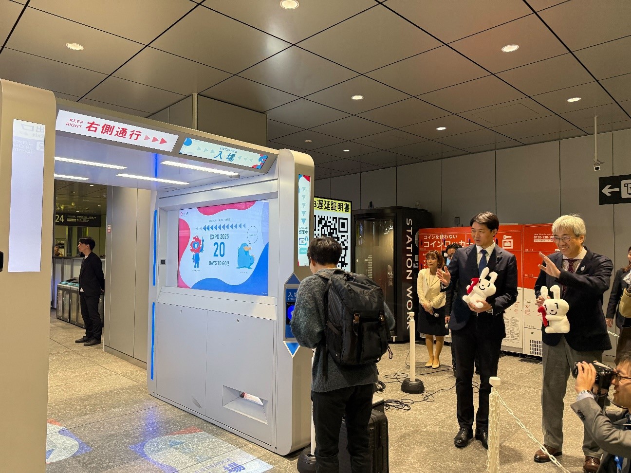 改札の右側にマイナちゃんのぬいぐるみを持った岸デジタル大臣政務官と西日本旅客鉄道株式会社の奥田本部長が立っており、学生が改札を通過する様子を見ている。