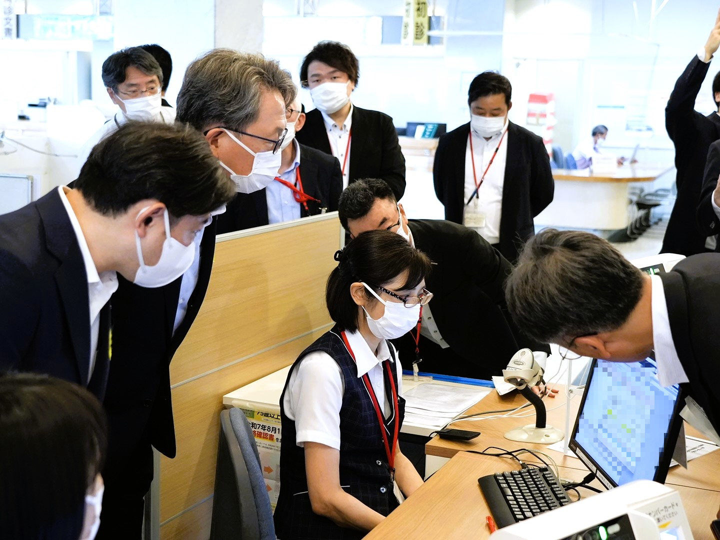 机の上にモニター画面が置いてあり、モニターの前には人が座っている。その後ろに平デジタル大臣と福岡厚生労働大臣が立ち、モニターを見ている写真