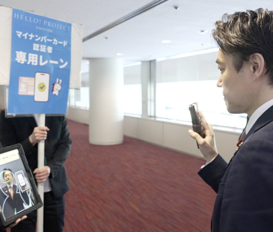 左側に「マイナンバーカード認証者専用レーン」と記載されている看板と、その手前に認証用のタブレットがあり、右側から穂坂デジタル副大臣がスマートフォンで二次元コードを表示しながら顔認証を実施している