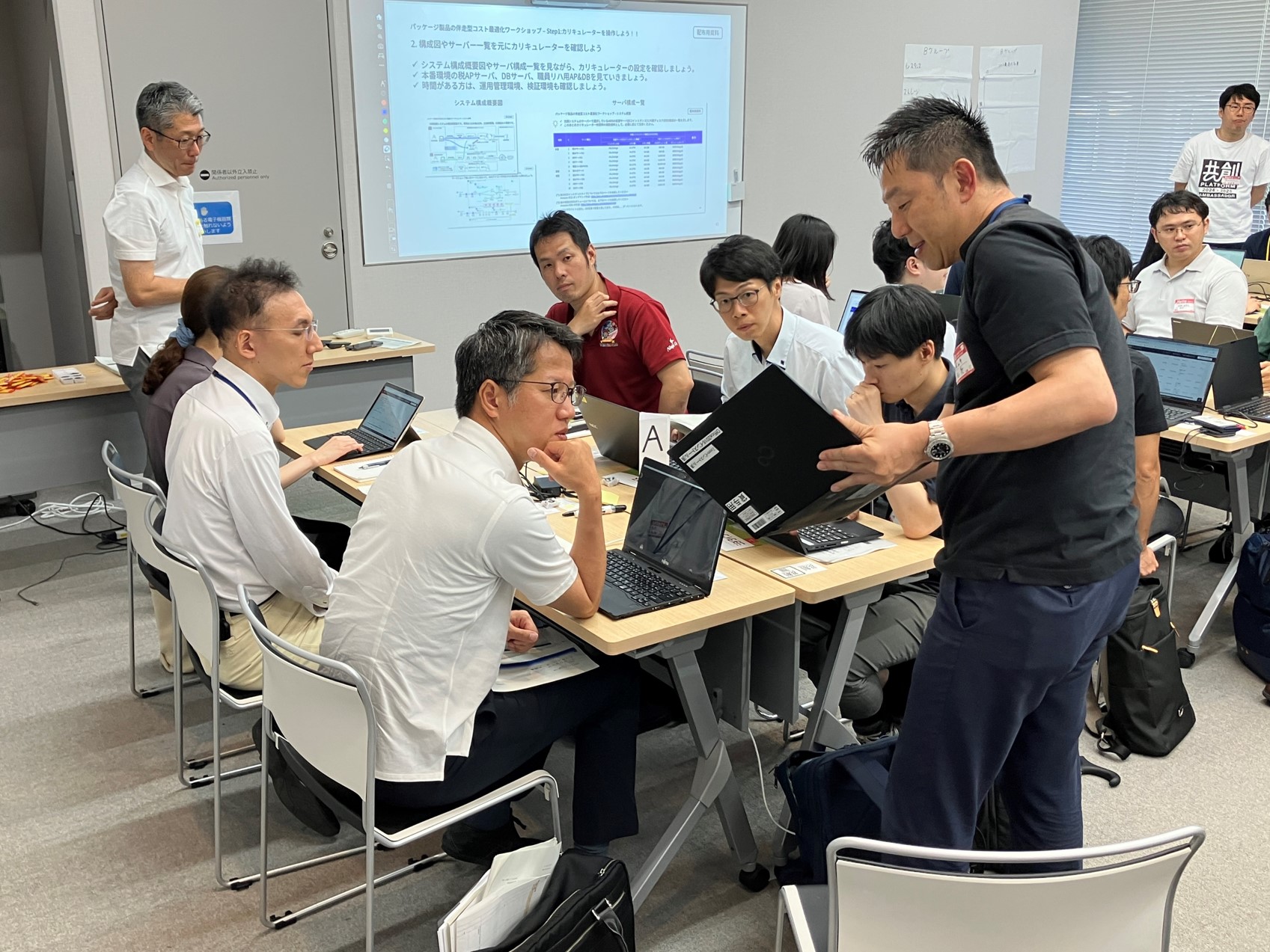 机を向かい合わせて6名が座っているところに、1名がパソコンの画面を見せながら立って説明している様子の写真。広い会議室で、隣にも同様の机と参加者が写っている。