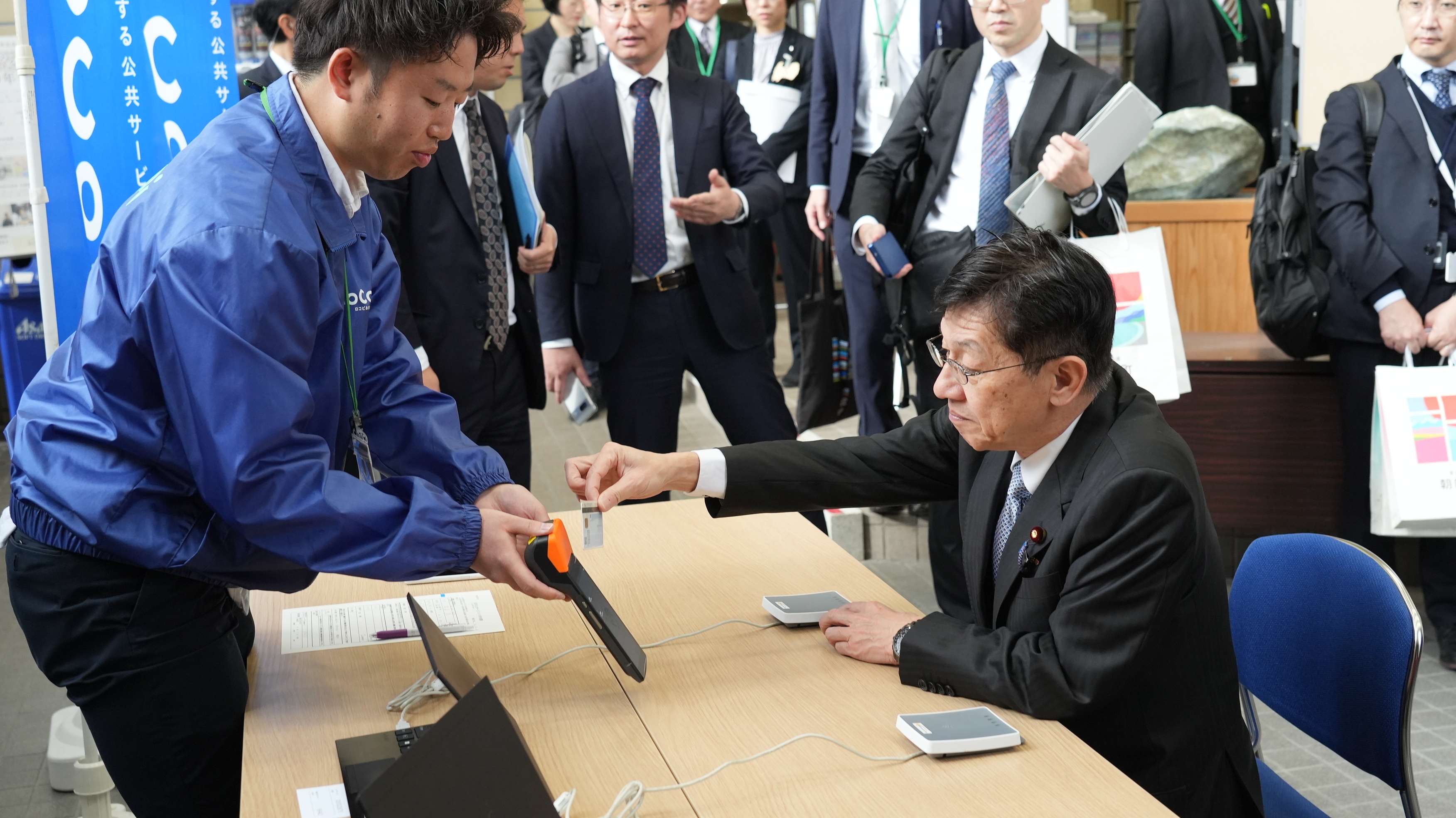 青いジャケットを着た職員の説明を受けながら、松本デジタル大臣が電子端末にマイナンバーカードをかざし、LoCoPiあさひまちの利用登録をする様子。