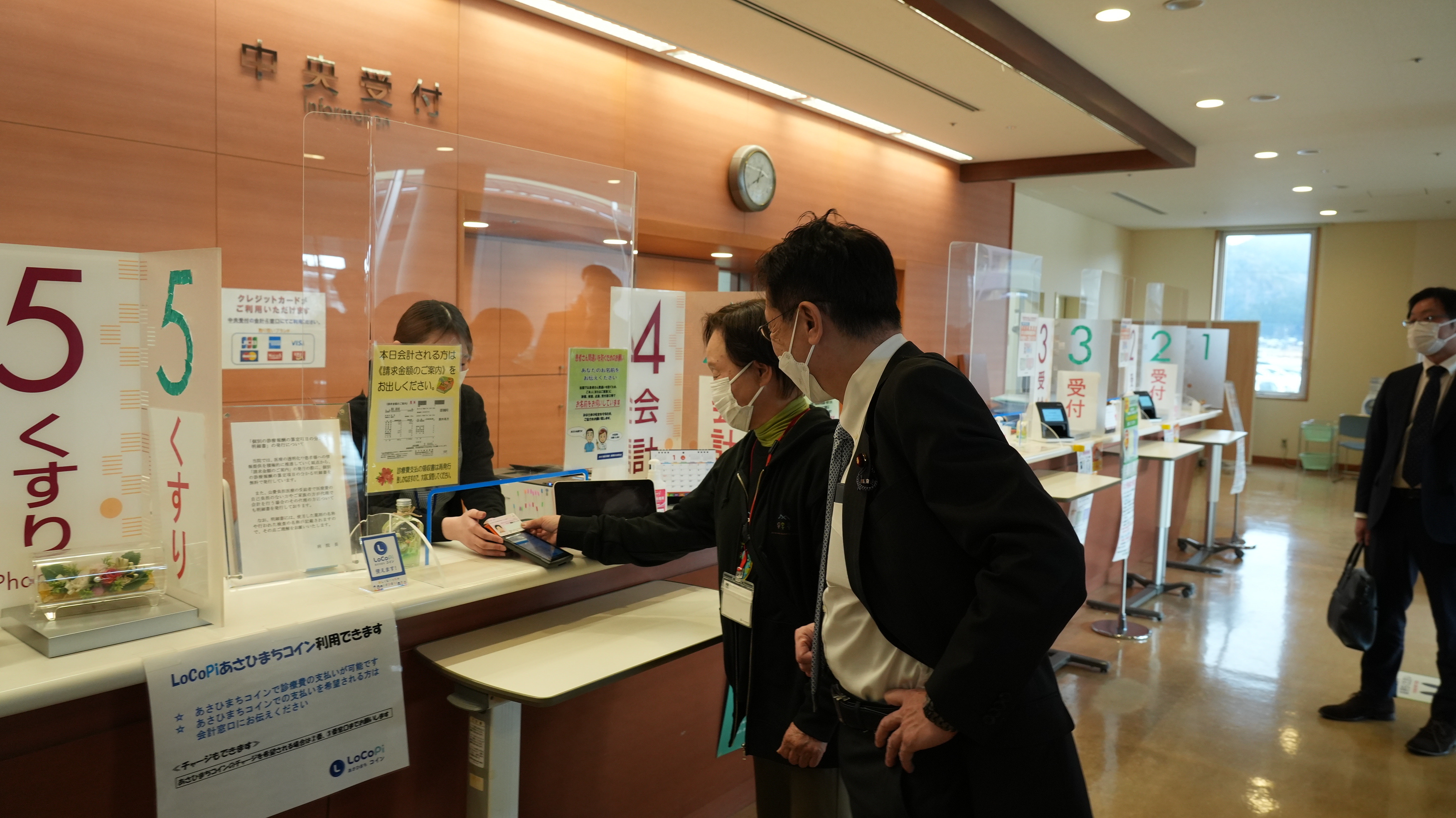 あさひ総合病院の受付で、病院利用者が専用端末にマイナンバーカードをかざしている様子を視察する松本デジタル大臣。