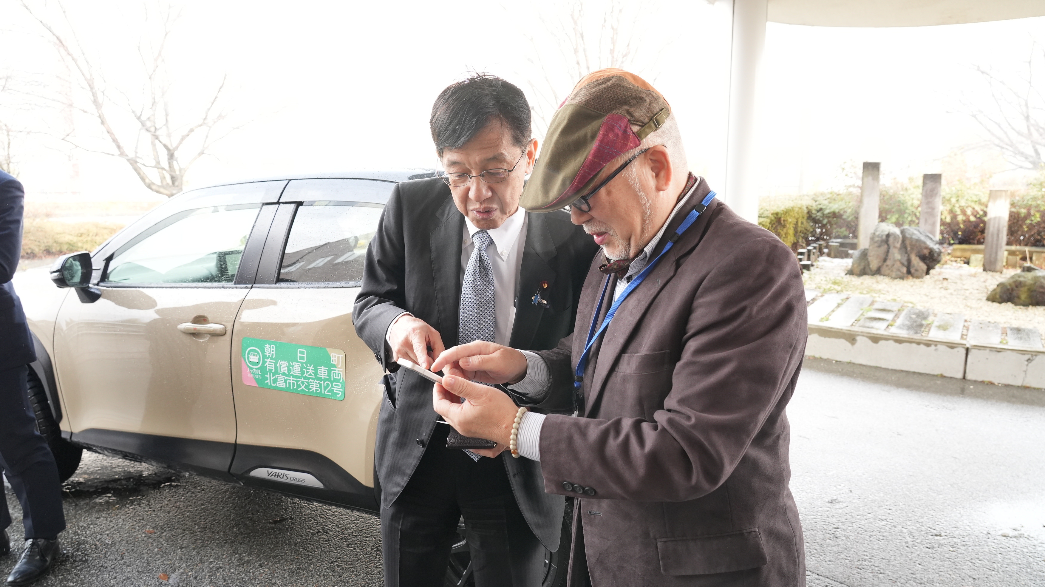 自動車の横で、松本デジタル大臣と男性がスマートフォンを操作しながら、まちバスチケットを使って決済をする様子。