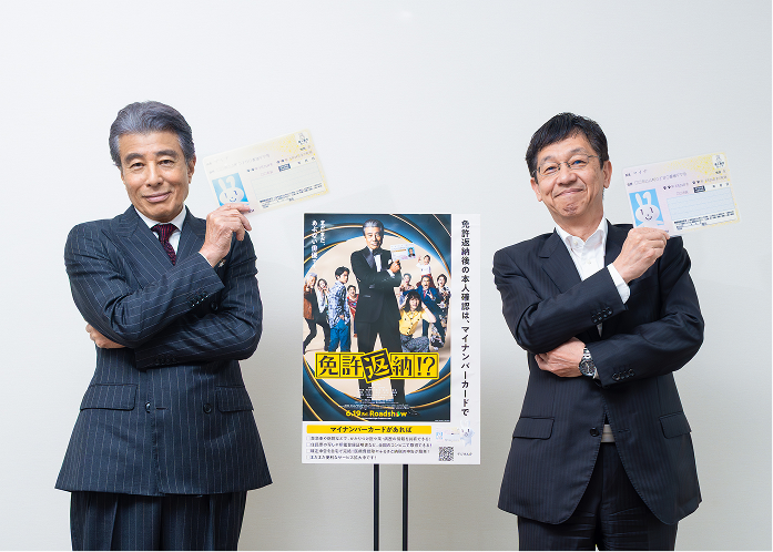 松本デジタル大臣と映画「免許返納!?」主演の舘ひろしさんが、スーツ姿で両腕を胸の前で組みながらマイナンバーカードを左肩の上に掲げ、中央に置かれた映画「免許返納!?」とマイナンバーカードのタイアップポスターに映る舘ひろしさんと同じポーズで、笑顔でカメラに向かっている。