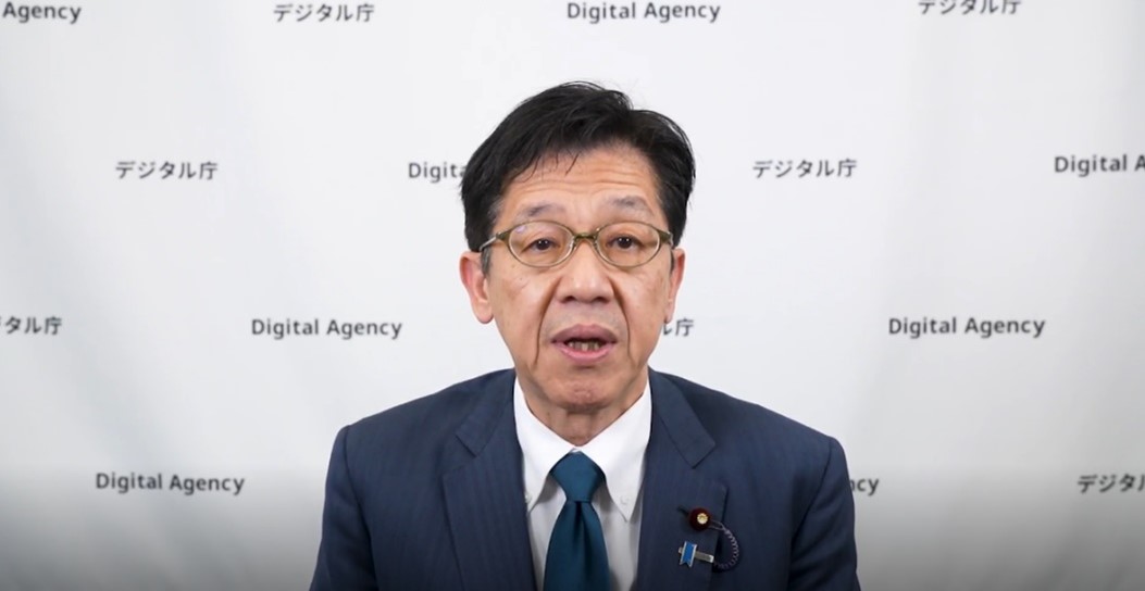 デジタル庁のロゴが繰り返し配置された白い背景の前で、ネイビースーツ姿の松本デジタル大臣が正面を向いてカメラに向かって発言している写真。