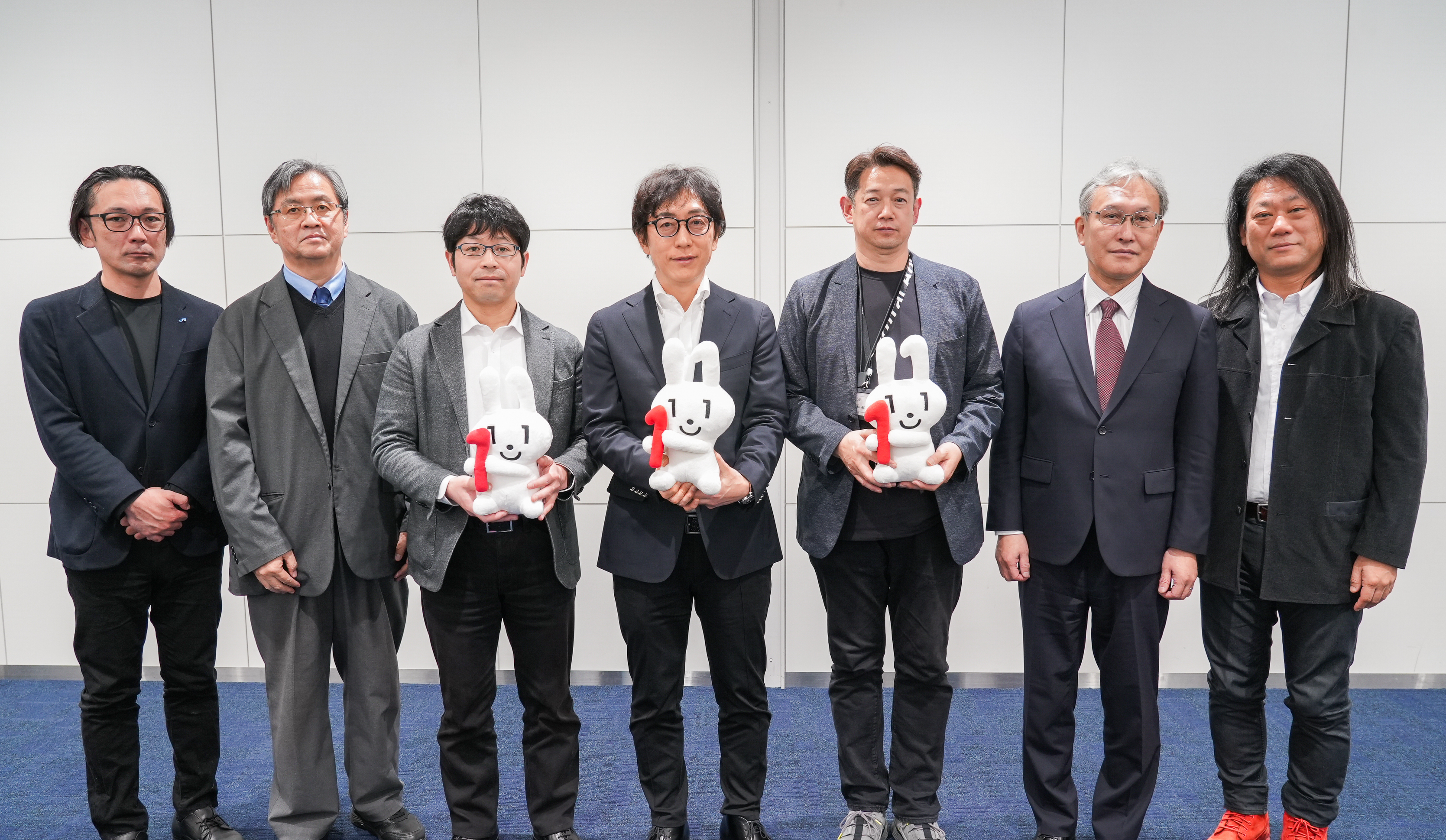シンポジウムにて参加者が記念撮影する様子。7名の男性が横一列に並んで記念撮影している。左から西日本旅客鉄道株式会社 眞鍋宗一郎課長、順天堂大学 満塩尚文准教授、デジタル庁国民向けサービスグループ 久芳全晴企画官、デジタル庁国民向けサービスグループ 鳥山高典企画調整官、デジタル庁国民向けサービスグループ 小畑雅人参事官補佐、国立情報学研究所 佐藤周行教授、一般社団法人OpenIDファウンデーションジャパン 富士榮尚寛代表理事が立っている。中央の3名が赤い「1」の数字が描かれたマイナちゃんのぬいぐるみを手に持っている。