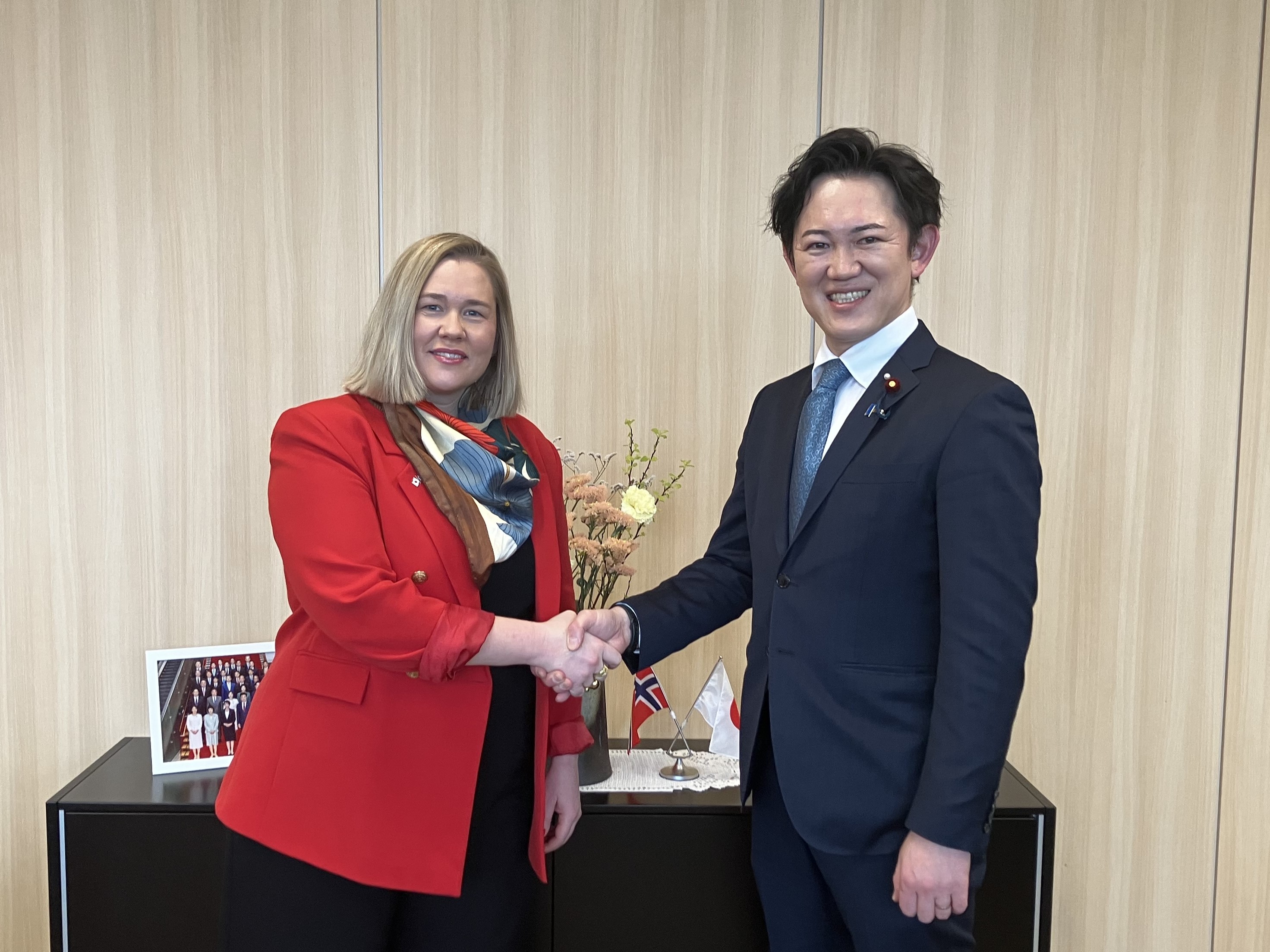 Ms. Marte Ingul, acting state secretary to Minister of Digitalisation and Public Governance, Kingdom of Norway on the left and the Parliamentary Vice-Minister for Digital Transformation Kawasaki on the right, standing in front of a light wood-paneled wall, smiling as they shake hands. Ms. Ingul wears a red blazer over a dark outfit with a patterned scarf, and Mr. Kawasaki wears a dark blue suit with a white shirt and tie. Behind them is a cabinet displaying small Norwegian and Japanese flags, a vase of flowers, and a framed group photo.
