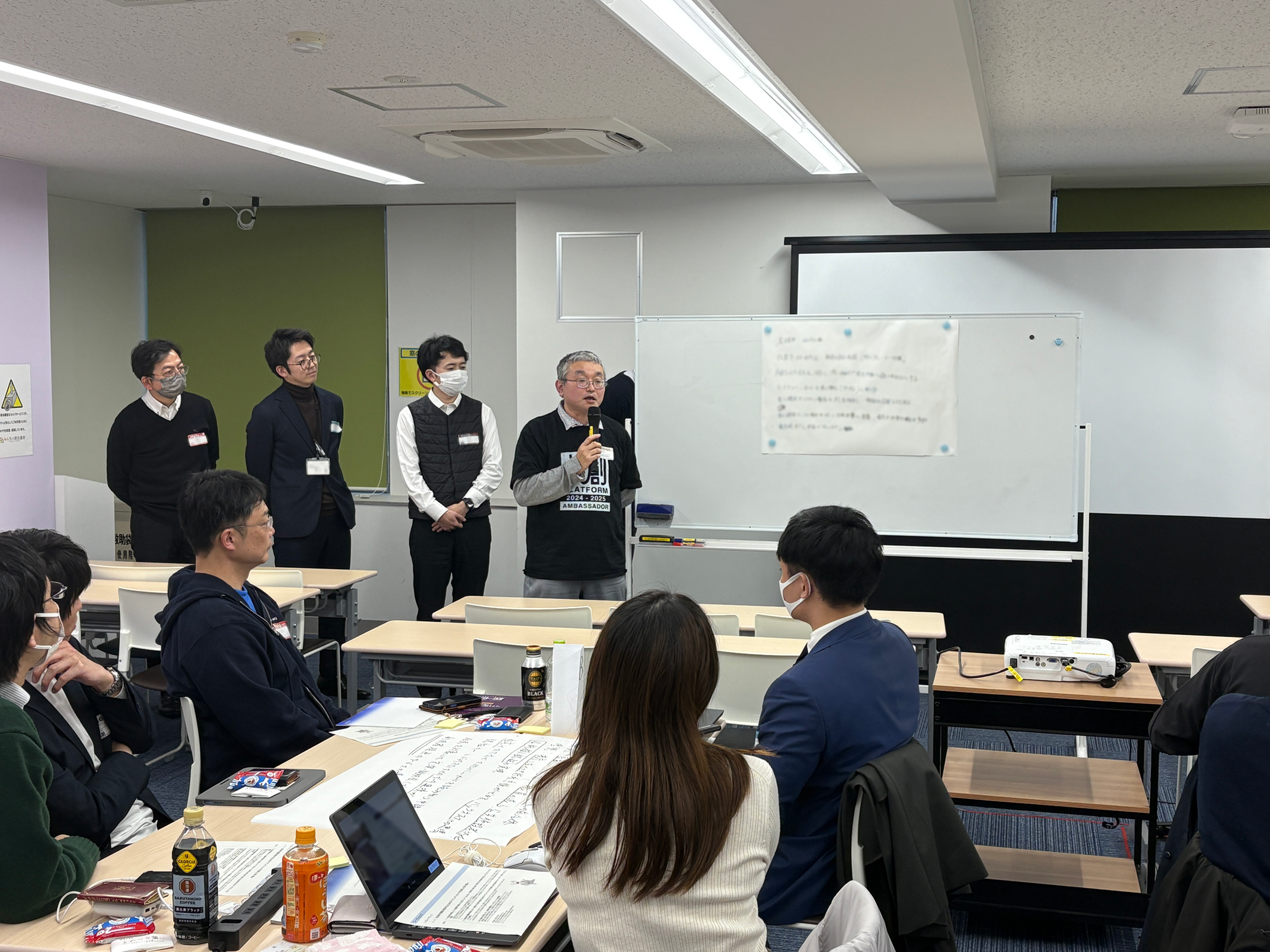 ワークショップの発表をしている写真。4名が模造紙を貼り付けたホワイトボードの横に立ち、そのうち1名がマイクを持って発表している。発表者の前には5名が1つの机を囲みながら座っており、発表者の方を向いて発表内容を聞いている様子。