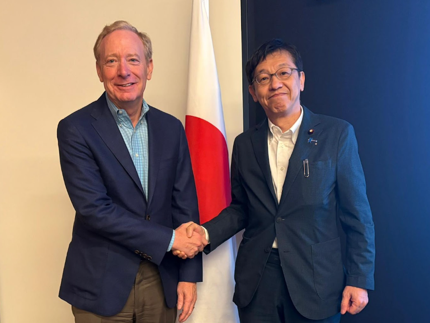 日本の国旗の前で、ブラッド・スミス副会長兼社長と松本デジタル大臣が握手をしている様子。