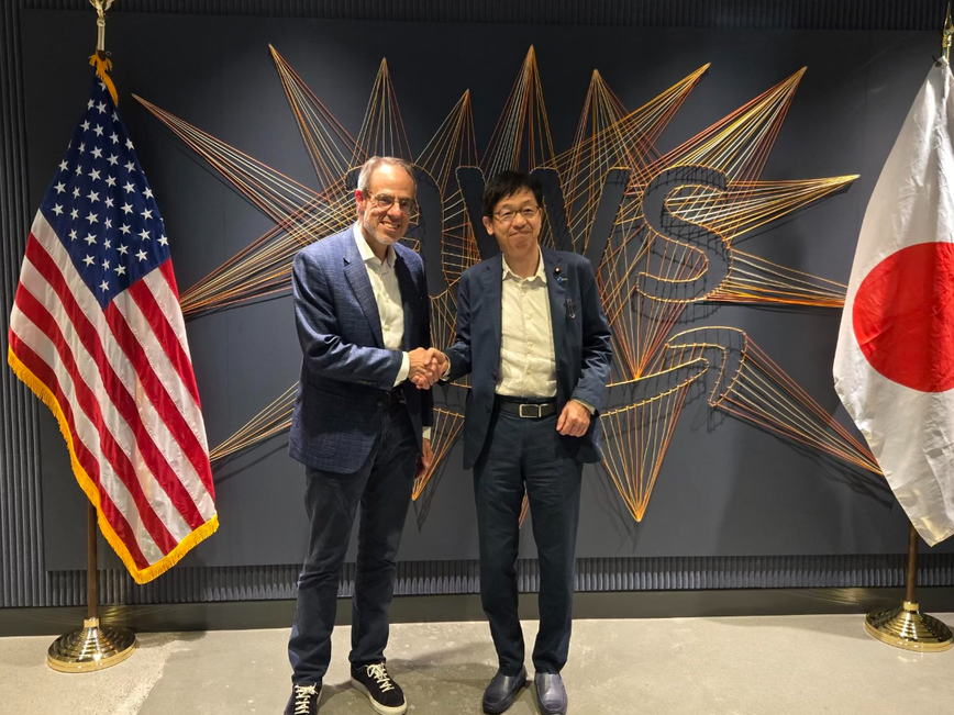 awsと書かれた壁の前に日米両国の国旗があり、左側にデイビッド・ザポルスキー上級副社長、右に松本デジタル大臣が立ち、握手をしている様子。