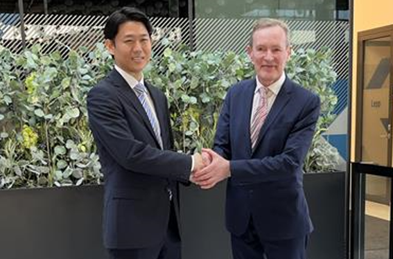 2人が室内で並んで立っており、握手を交わしている写真。背後には緑の植物が写っている