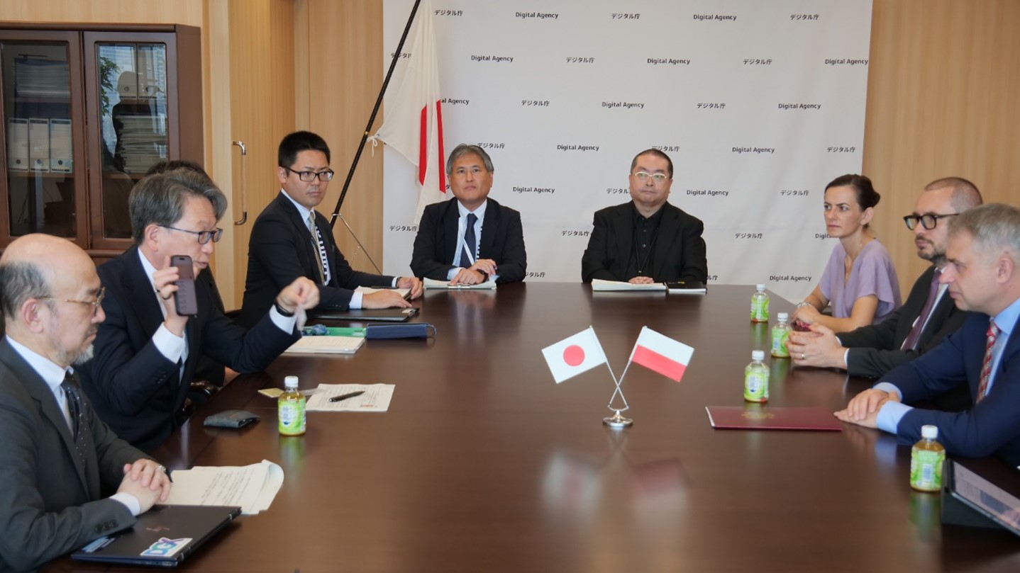 テーブルを挟んで左側に平大臣、右側にガフコフスキ副首相兼デジタル化大臣が座って会談している写真。平大臣は、右手にiPhoneのマイナンバーカードが表示されたスマートフォンを持ち、画面を見せながら説明している。テーブルの中央には、日本とポーランドの旗が写っている。