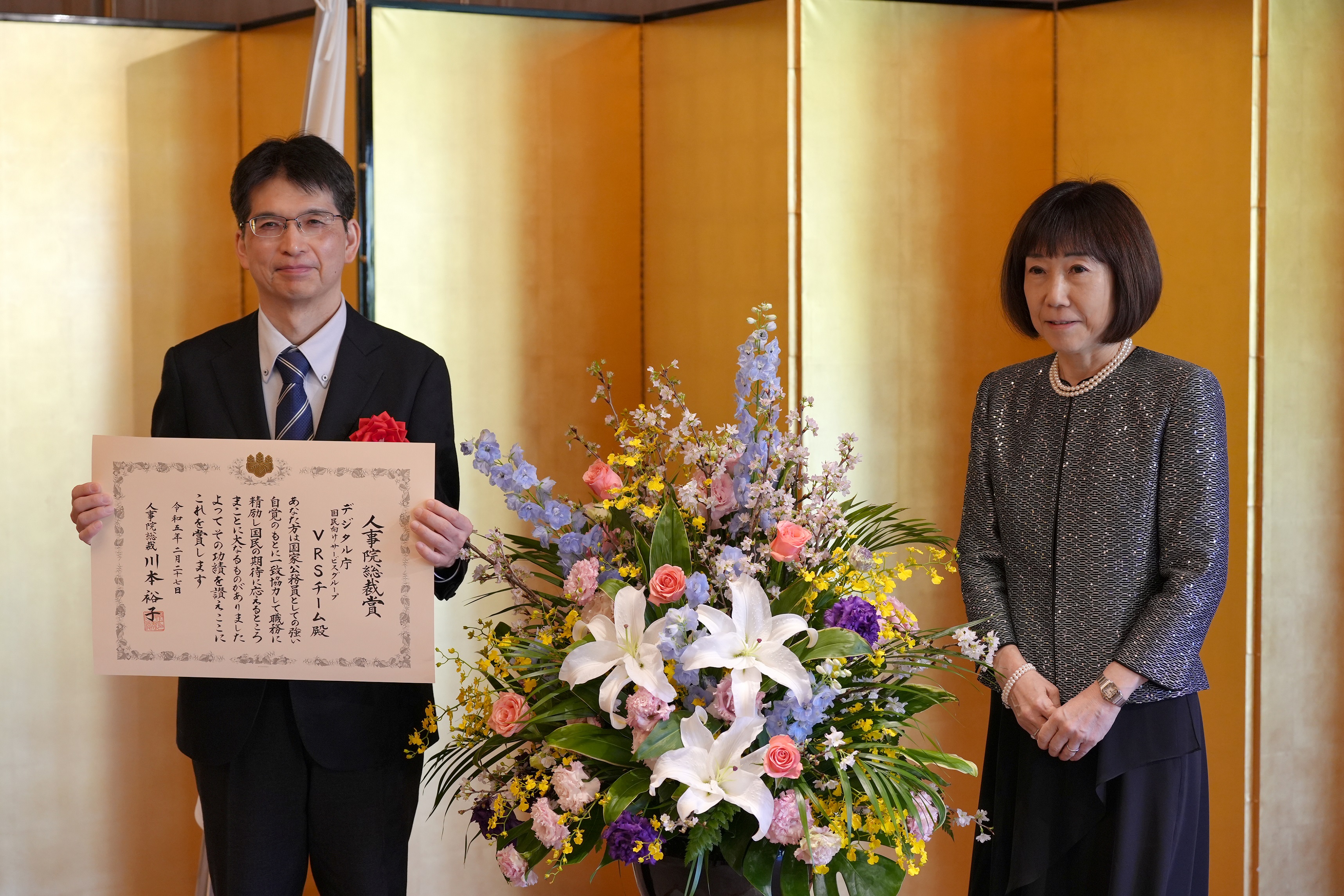 左から順番にデジタル統括官と川本裕子総裁(人事院)が立って並んでいる。統括官は表彰状を胸の前に広げ、両手で持っている。
