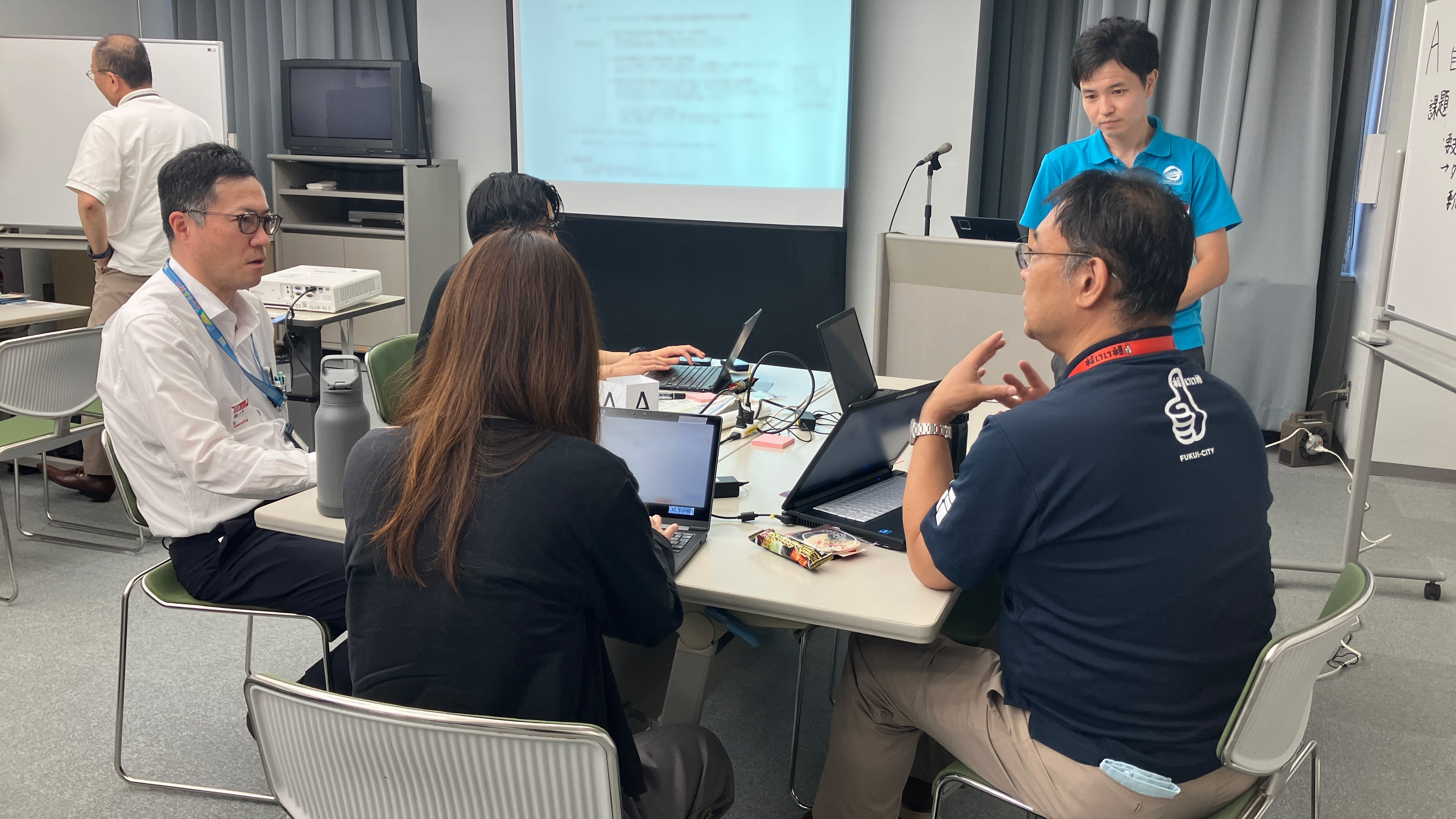 The photo shows four people sitting at desks facing each other. One person is standing around the desk and listening to the four people. Several people are talking while looking at the computer screen.