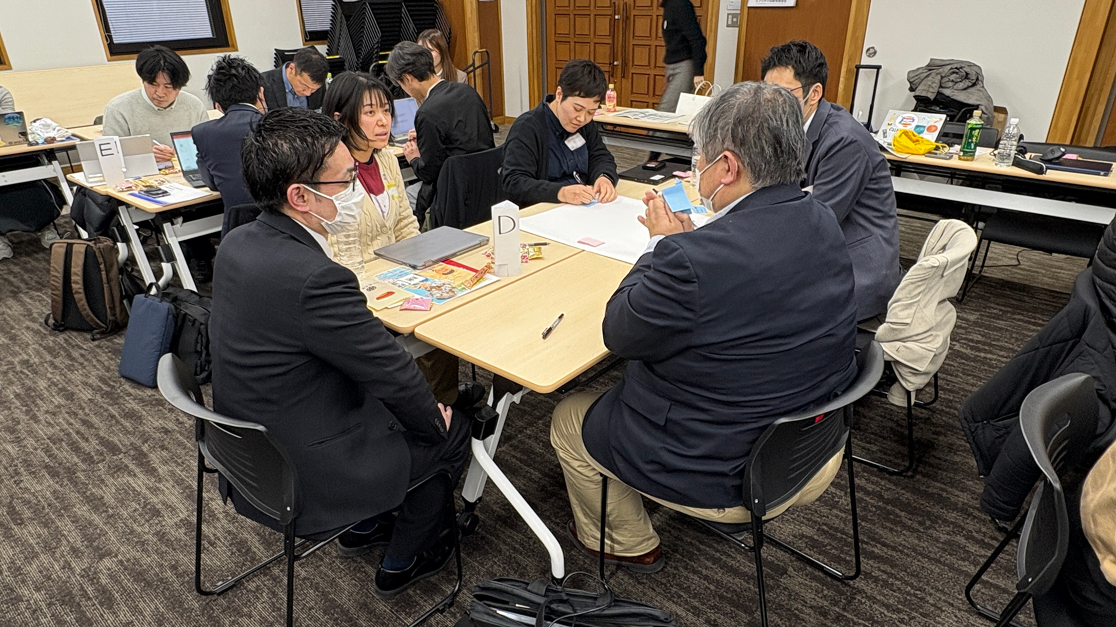 ワークショップの写真。机を囲んで5名が話し合っている様子。机の真ん中には模造紙が広げられており、１人が模造紙にメモを取っている。机の上には「D」と書かれた札が置かれている。