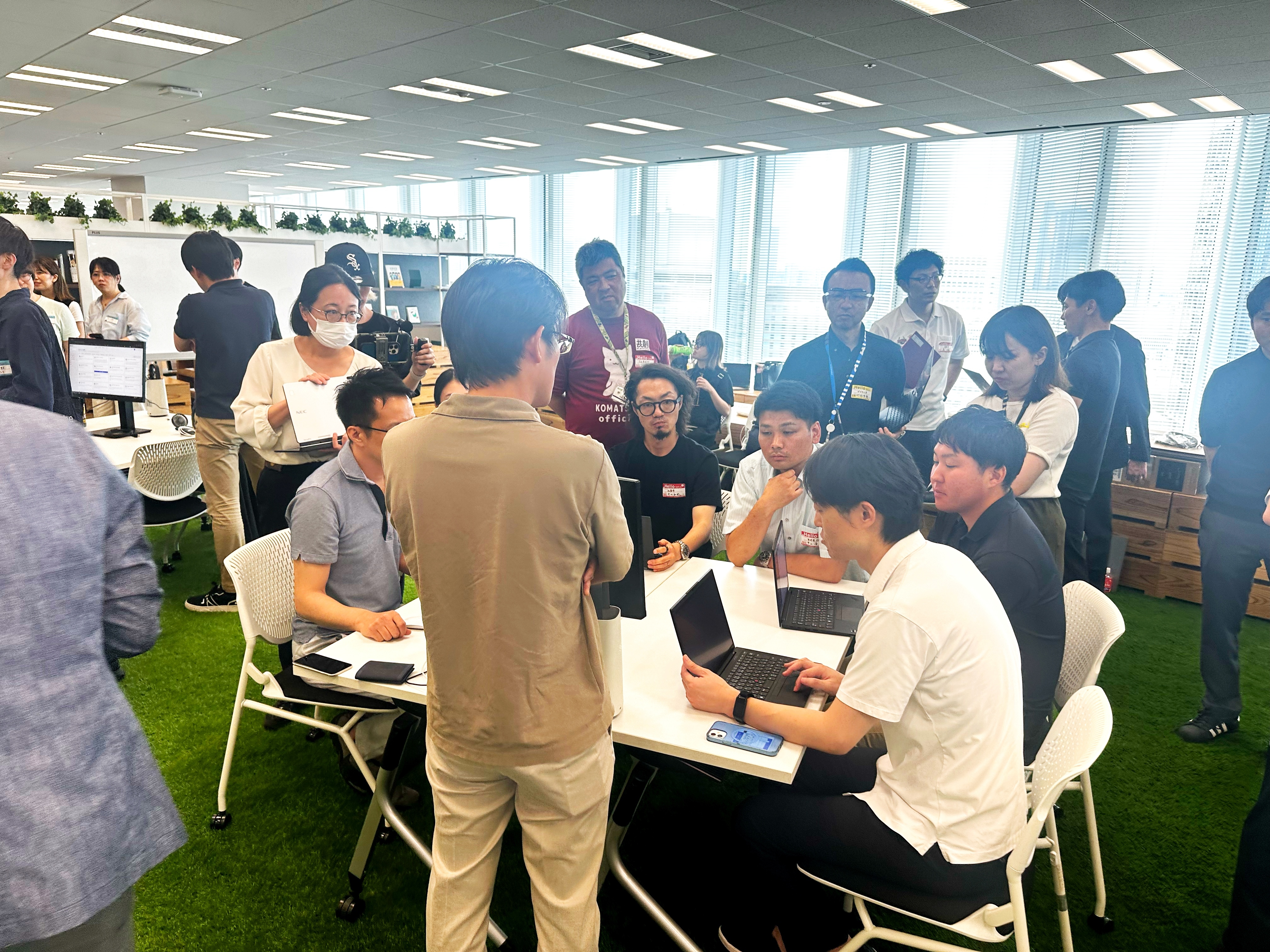 1つの机に対して、参加者6名と事務局担当者4名が、1人の説明者の話を聞いている。後方や左右にも別の2〜3つのグループが机ごとに集まっている。