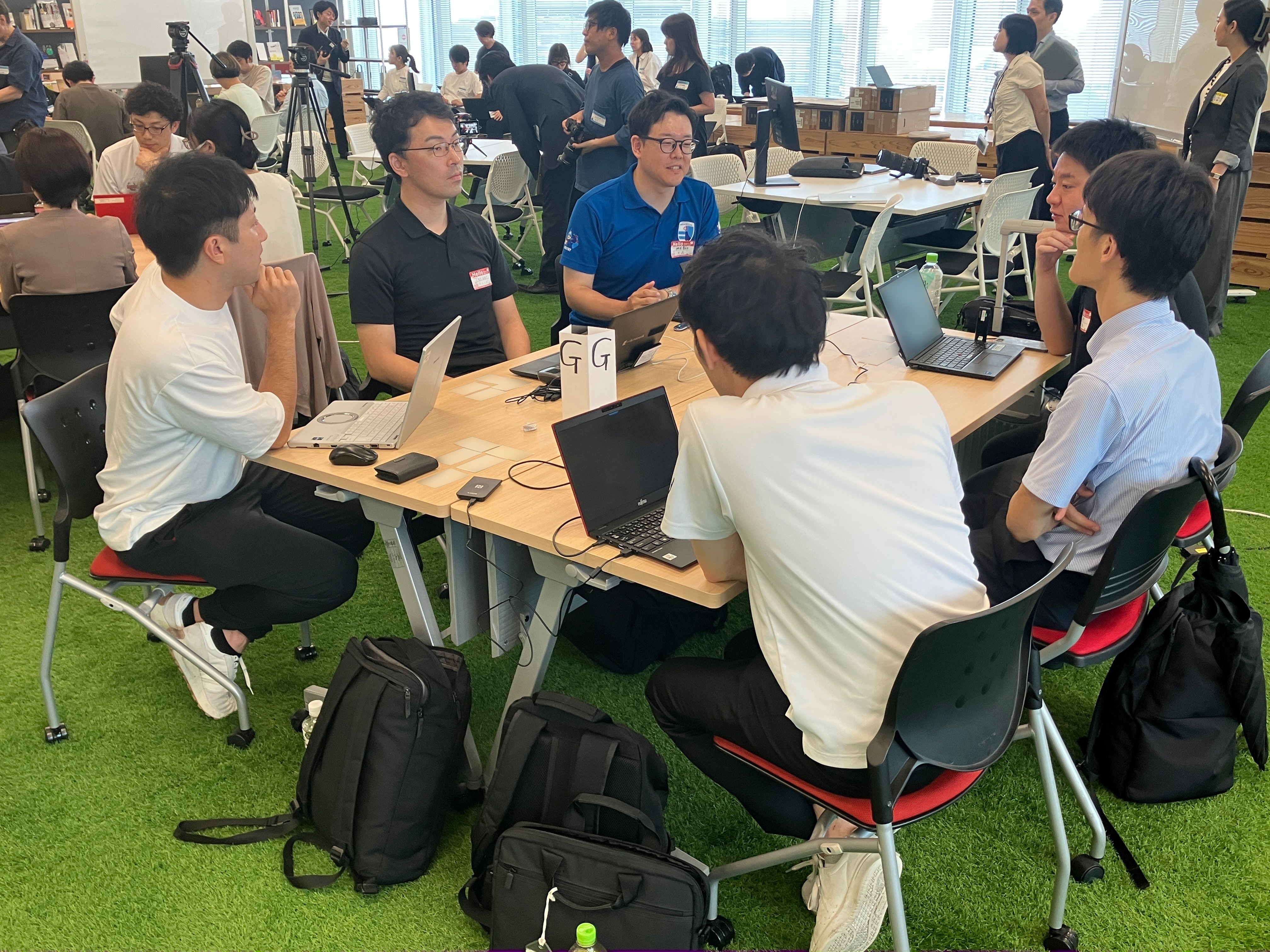 参加者6名が、自治体業務のAI活用について、ディスカッションしている写真。机の上には、「G」と書かれたグループ名を表す札が置かれている。後方や左右にも別の2〜3つのグループが机ごとに集まっている。