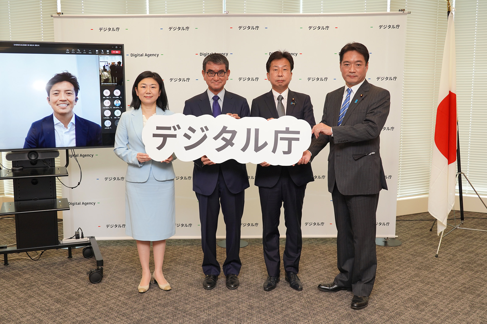 A group photo of the new and old government officials. From the left, they are lined up with former Senior Vice-Minister KOBAYASHI (shown on the screen for remote participation), Minister Dozen MAKI, new Minister Kono, new Senior Vice-Minister Okushi, and new Parliamentary Secretary Ozaki. The other four hold the Digital Agency logo in their hands.
