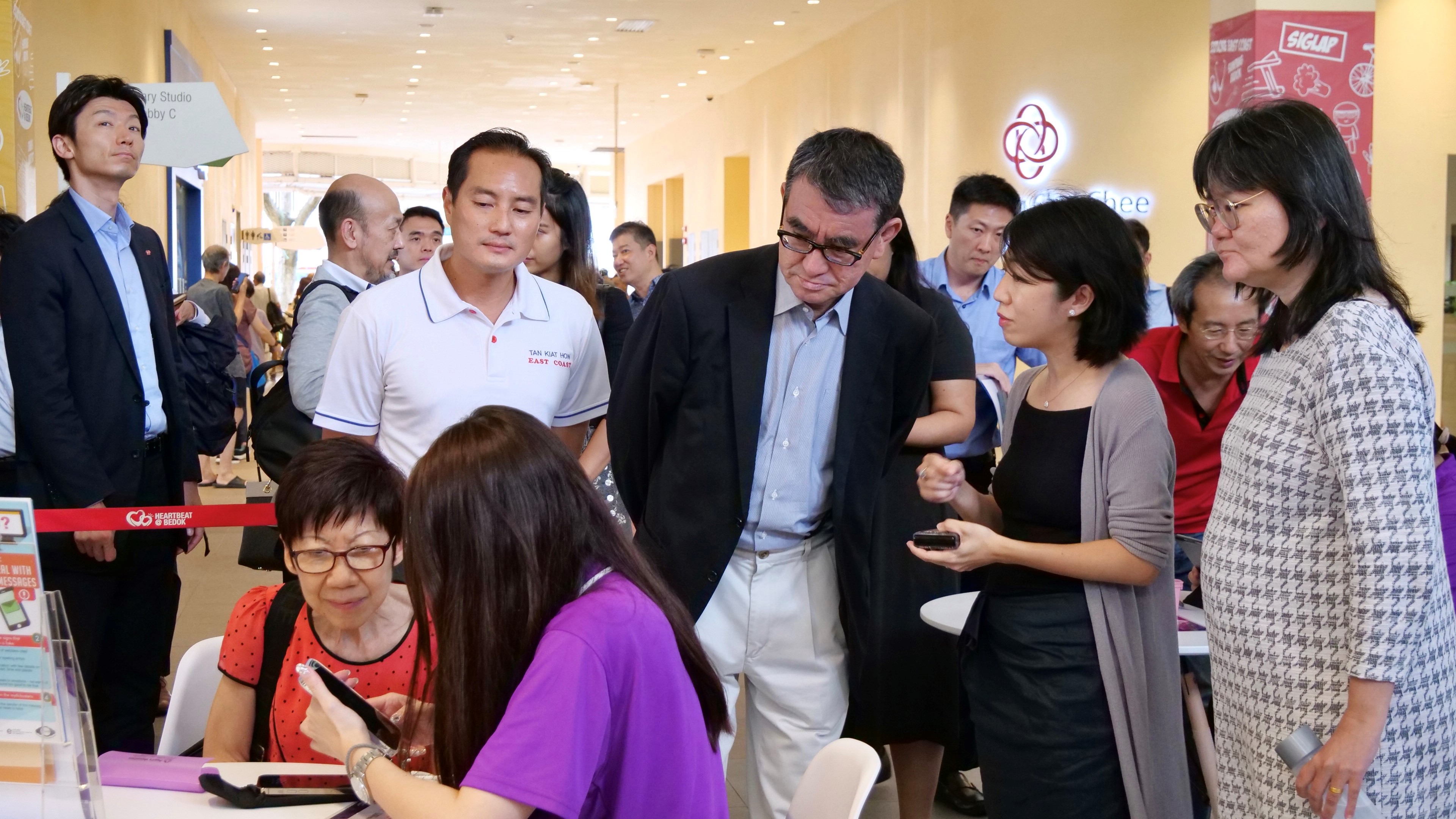 A photo captures Minister Kono observing a Digital Ambassador teach an elderly woman how to use a smartphone.