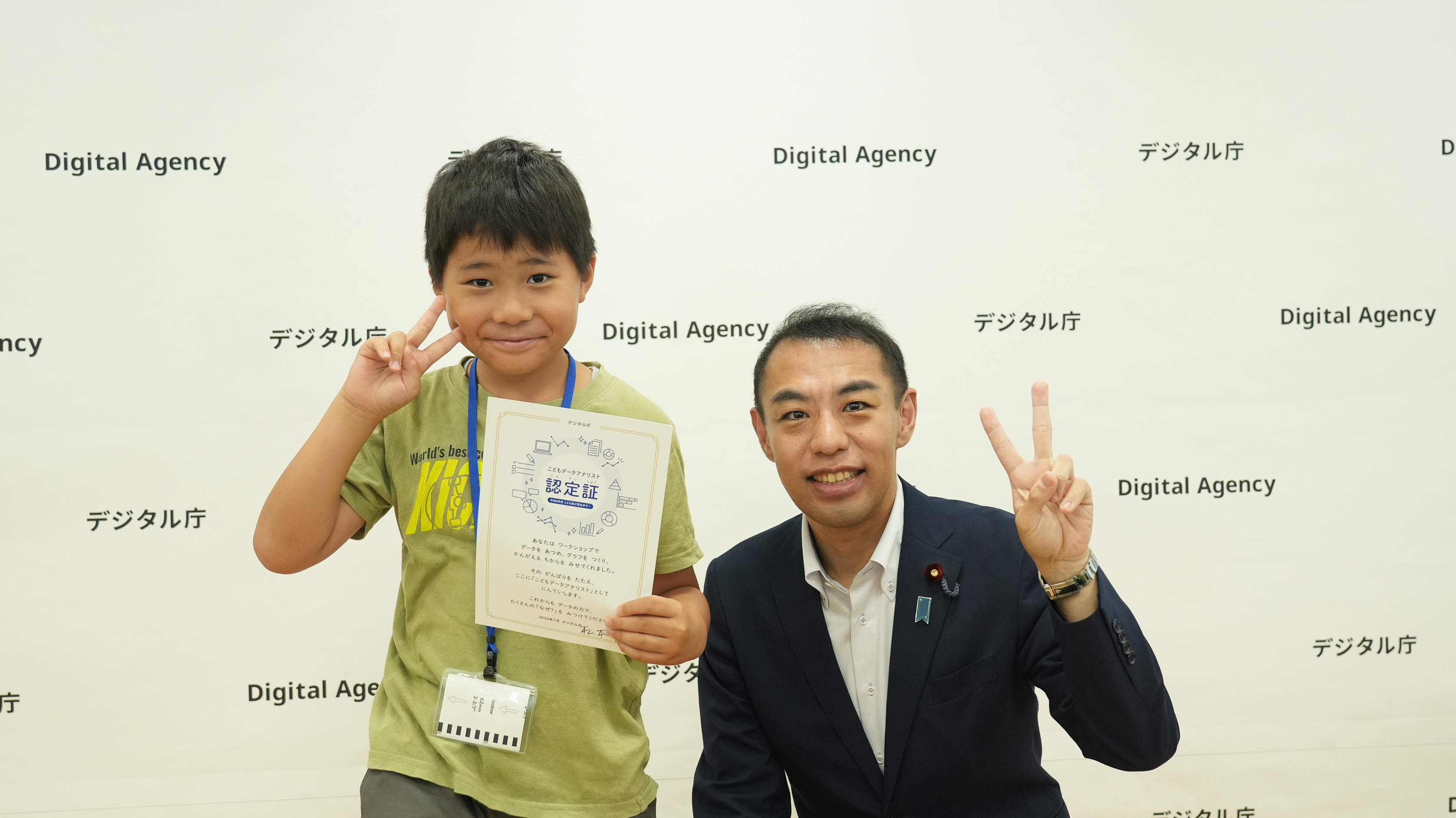 デジタル庁のバックパネルの前で、「こどもデータアナリスト認定証」を手にした男児と、副大臣がピースをして微笑んでいる記念写真