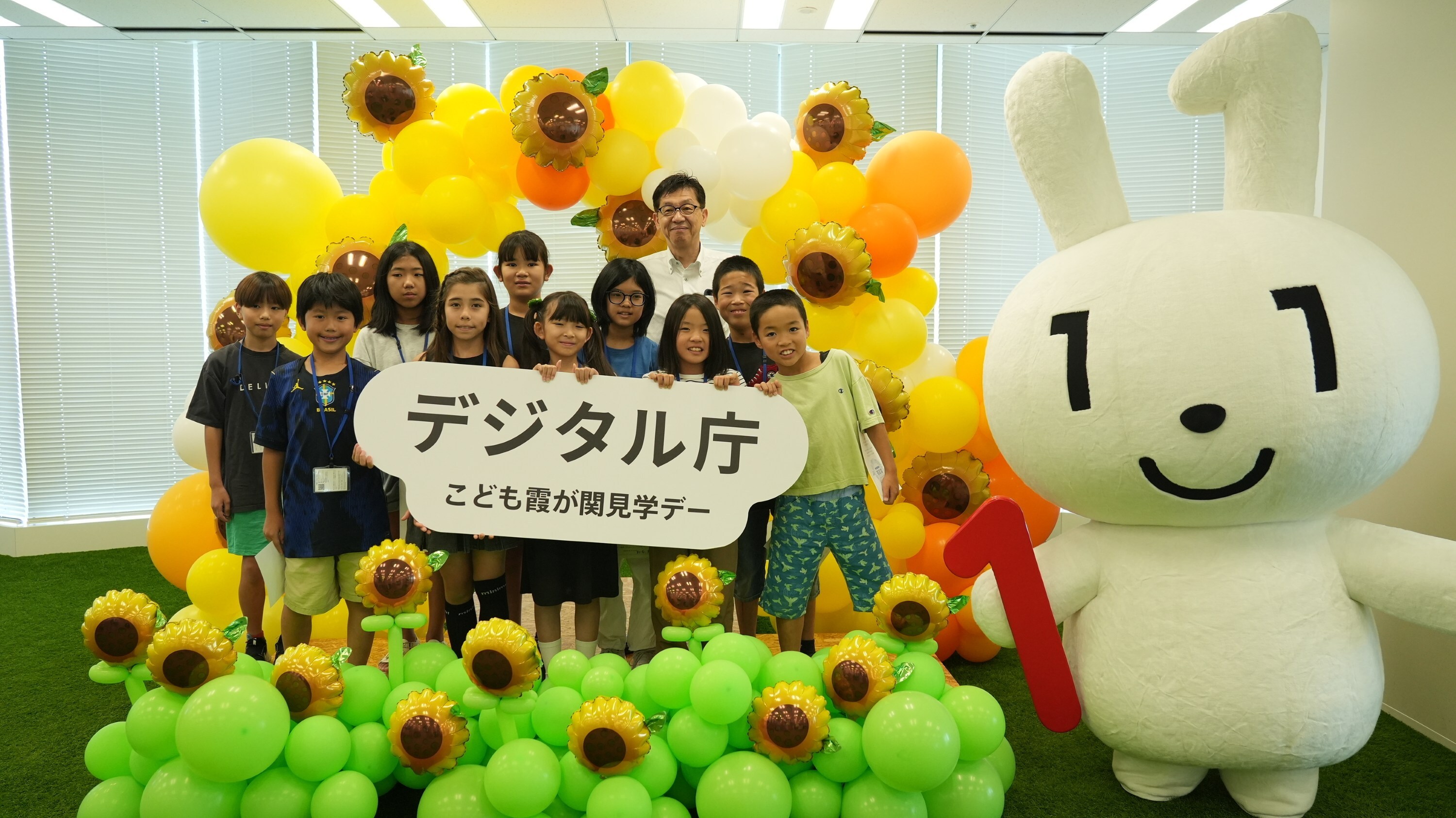 花のバルーンが飾られているフォトスポットに、子どもたち、大臣、マイナちゃんが並んで立っている集合写真