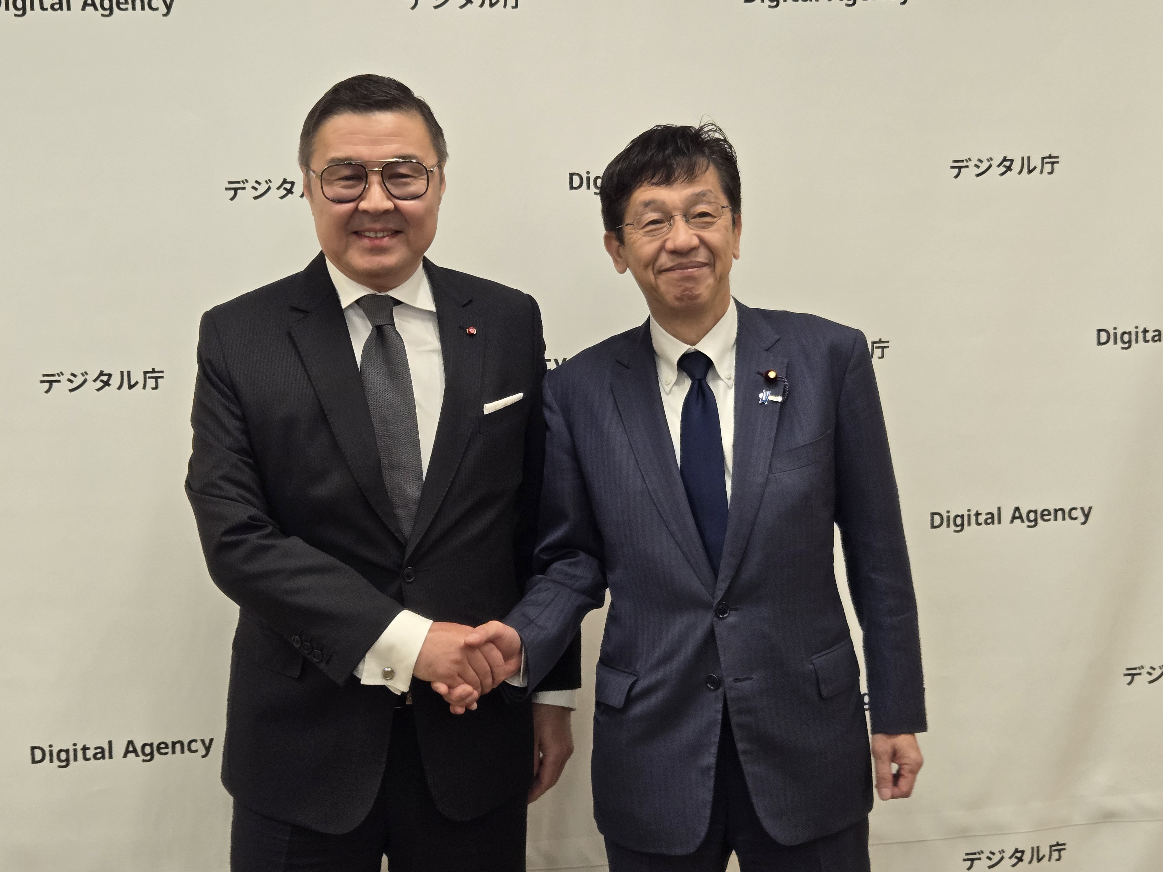 Minister for Digital Transformation Matsumoto is on the right, shaking hands with Mr. Yerlan Baudarbek-Kozhatayev, the Ambassador Extraordinary and Plenipotentiary of the Republic of Kazakhstan to Japan, both smiling. In the background is the Digital Agency's backdrop with logo mark.