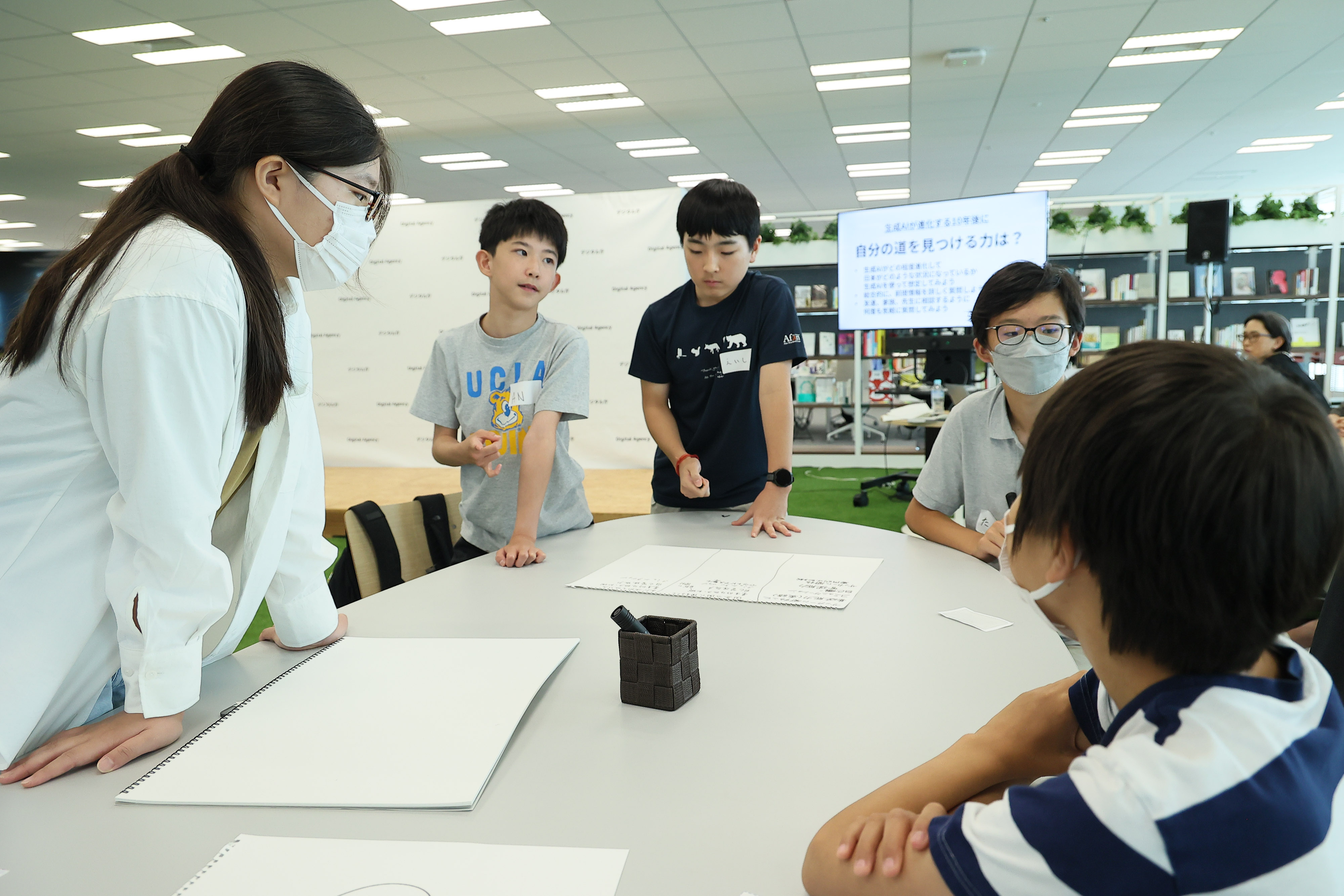 ワークショップ参加者の中学生5名で机を囲んで議論している。机の上にスケッチブック３つと黒色のペン1本が置いてある。背後のモニターには、議論のテーマが映し出されており、「 自分の道を見つける力は？」と記載されている。