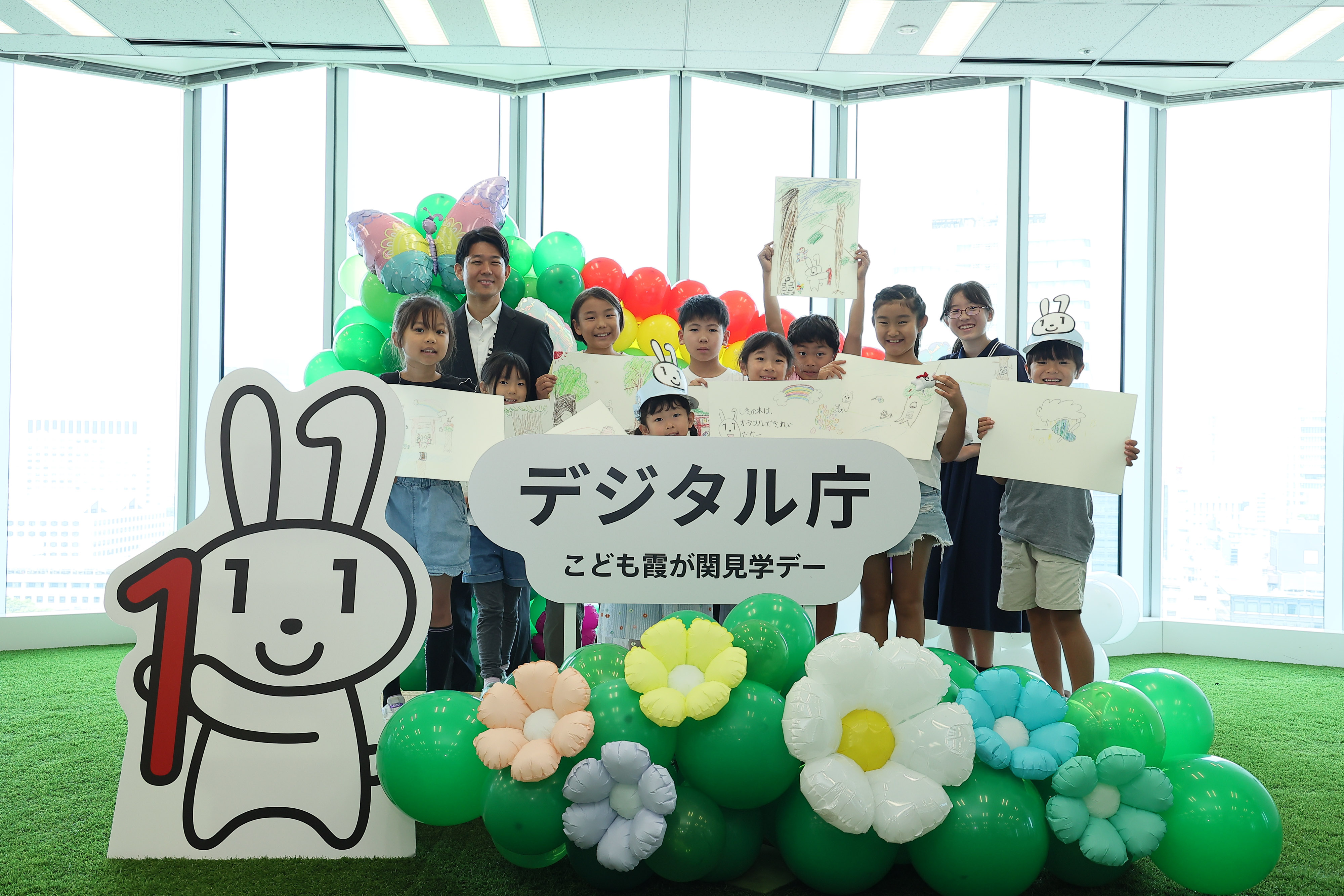 「デジタル庁 子ども霞が関見学デー」のロゴパネル、マイナちゃんのパネル、花や蝶のバルーンが飾られているフォトスポットに、ワークショップに参加した小学生と岸デジタル大臣政務官が立っている。小学生は、生成AIをつかって考えた「マイナちゃんの大冒険」の一場面を描いた画用紙を掲げている。​
