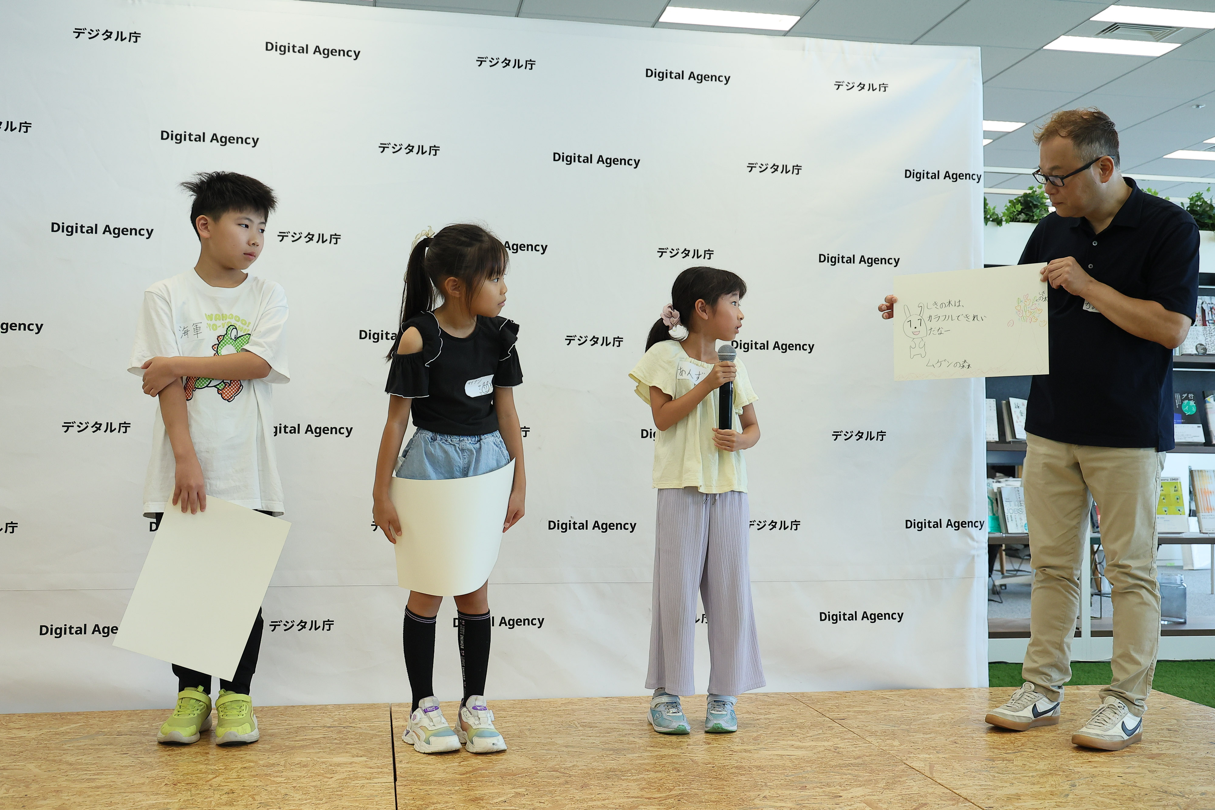 ワークショップ参加者の小学生3名が、デジタル庁のロゴをあしらったパネルが設置されたステージにたっている。​デジタル庁職員が右側に立ち、小学生のうち1名が描いた「マイナちゃんの大冒険」物語の絵を手に持っている。その小学生は、マイクを持ち絵の説明をしている。​小学生2名は、描いた絵を手に持ち発表されている絵を見ている。​