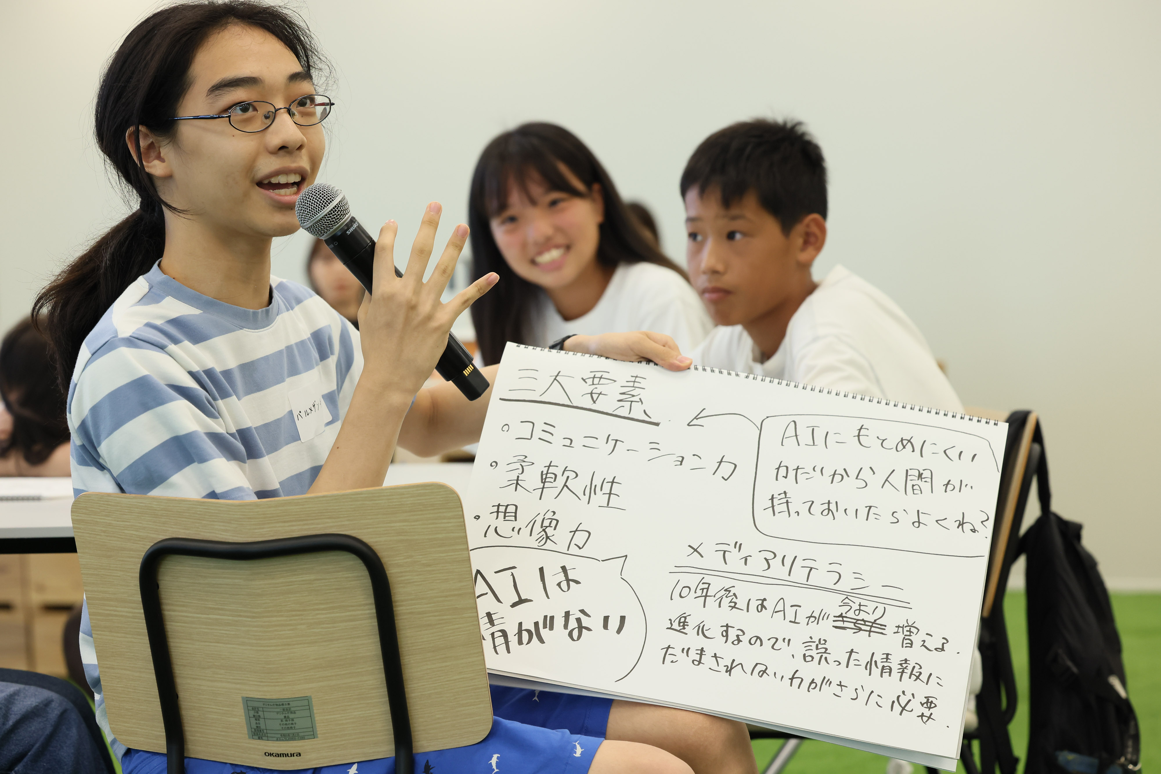 ワークショップ参加者の中学生3名のうち、1名が議論の内容をメモにまとめたスケッチブックを手に持ちマイクで発表している。​中学生2名は、スケッチブックを手に持って発表している中学生を見ている。