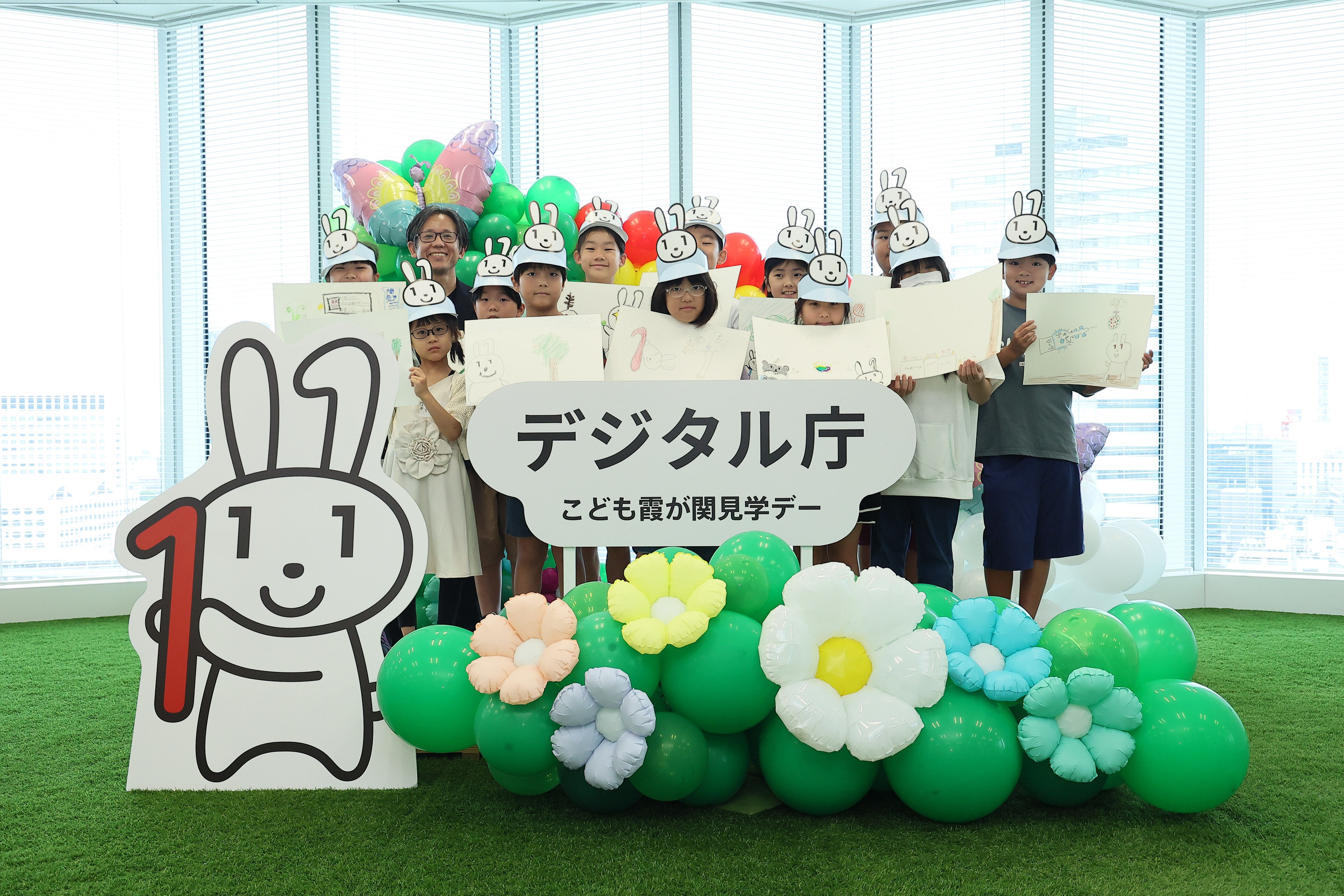 「デジタル庁 子ども霞が関見学デー」のロゴパネル、マイナちゃんのパネル、花や蝶のバルーンが飾られているフォトスポットに、ワークショップに参加した小学生と浅沼デジタル監が立っている。小学生は、生成AIをつかって考えた「マイナちゃんの大冒険」の一場面を描いた画用紙を掲げている。