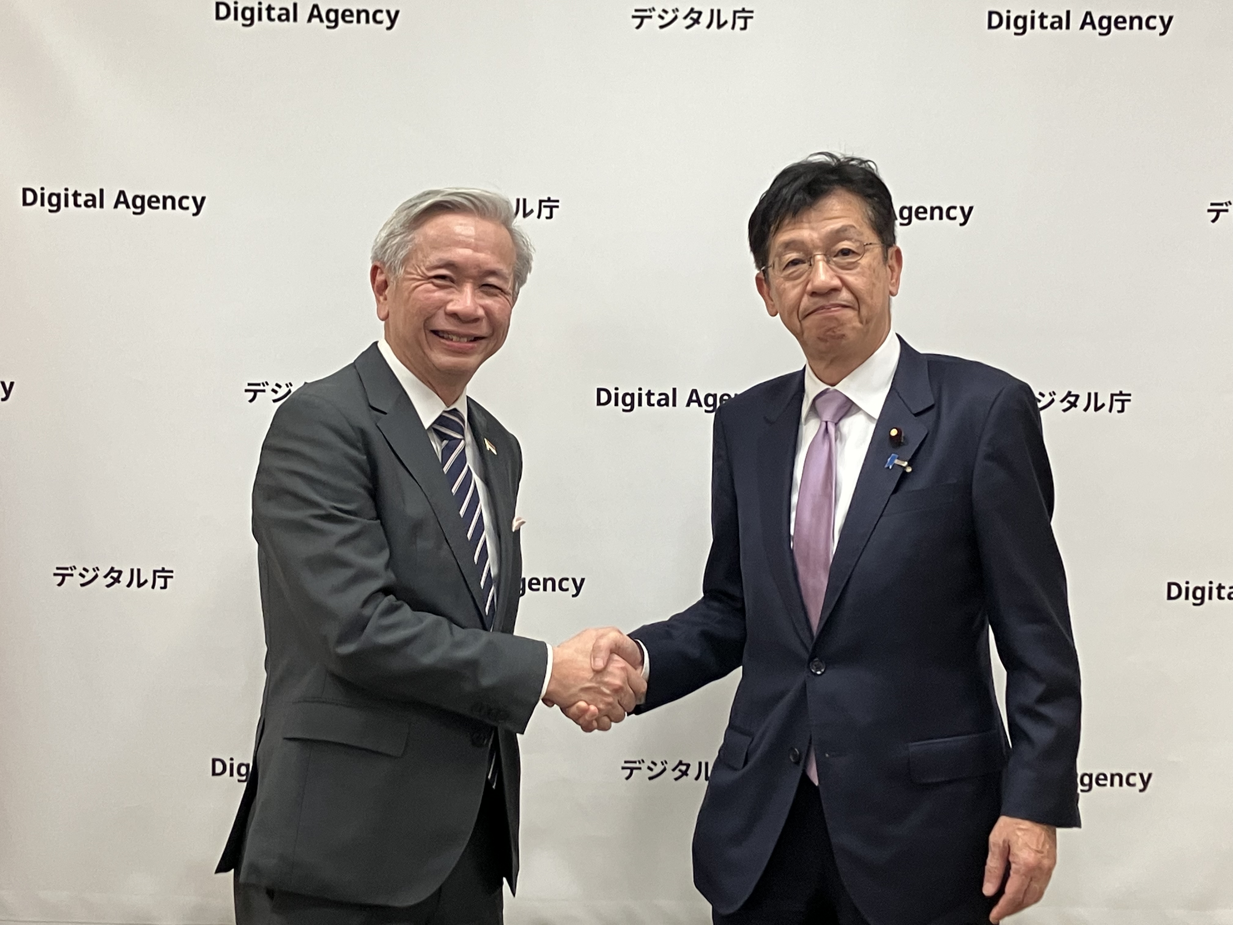 Minister for Digital Transformation Matsumoto (right) and Chief Executive Koh of Cyber Security Agency of Singapore (left) shake hands and smile, with the Digital Agency’s logo on the backdrop behind them.