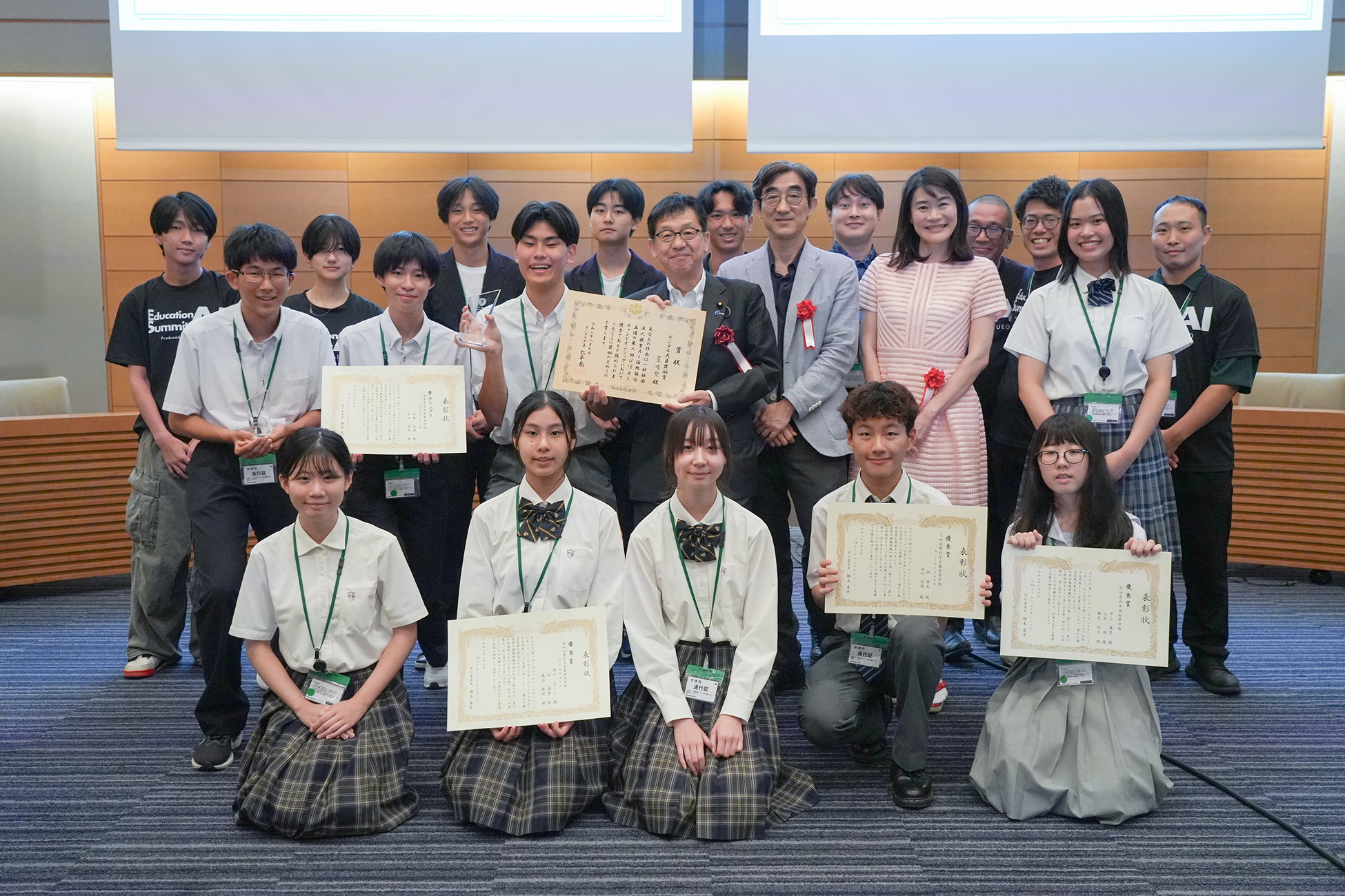 会議室にて、約20名が3列に並んで撮影した表彰式の集合写真。前列は学校制服姿の女子学生4名が床に座り、うち複数名が「表彰状」と記された賞状を手にしている。中列には参加学生が複数名が並び、中央の松本デジタル大臣がスーツ姿で赤いコサージュをつけ、大きな賞状を掲げている。背景は木目調のパネルが施された格式ある室内。