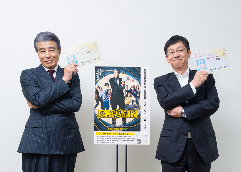 Minister for Digital Transformation Matsumoto and the lead actor Hiroshi Tachi stand side by side in suits, smiling with their arms crossed. Each holds a My Number Card above their left shoulder, copying Hiroshi Tachi's pose from the "Goodbye My Car" poster shown in the center.