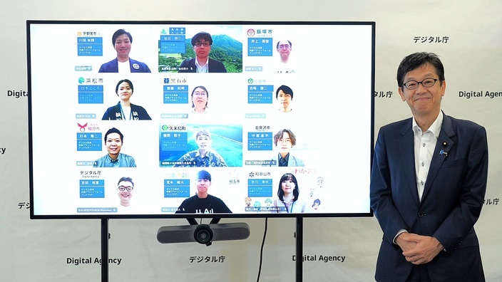 Minister for Digital Transformation Matsumoto stands beside a large screen in the Digital Agency office displaying the online inauguration ceremony for Digital Reform Co-Creation Platform Ambassadors.