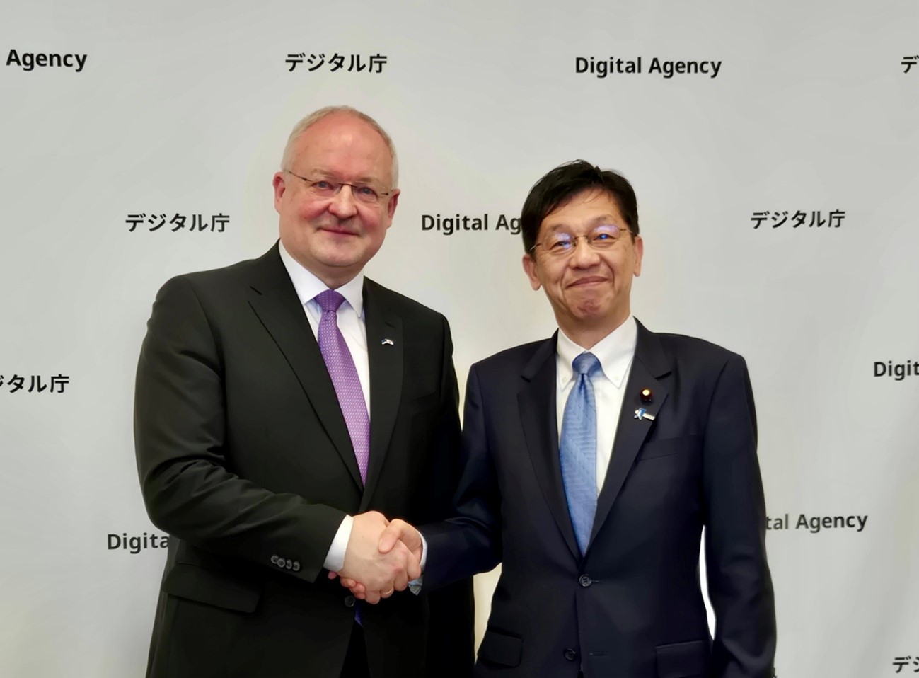 デジタル庁のロゴが繰り返し配置された白い背景の前で、左側にアンドレス・ウンガ駐日エストニア共和国大使、右側に松本デジタル大臣が正面を向いて立っている。二人は中央で笑顔で握手を交わしている。