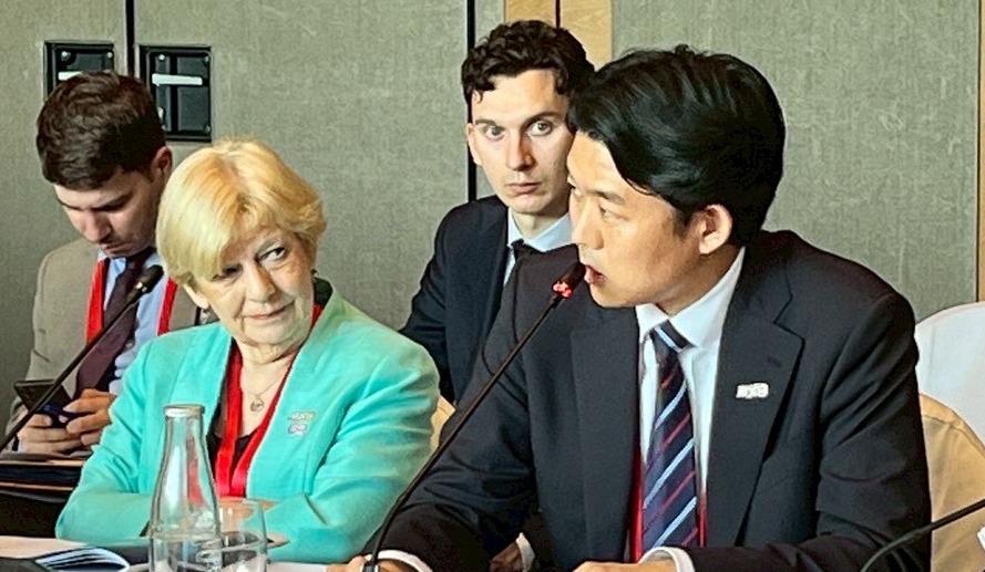 訪問先で会議中の写真。テーブルに着席した複数の参加者が映っている。左側には緑色のジャケットを着た人物が座っており、その右隣で濃紺のスーツを着た岸デジタル大臣政務官が発言している。テーブルの前には参加者の名札が置かれている。
