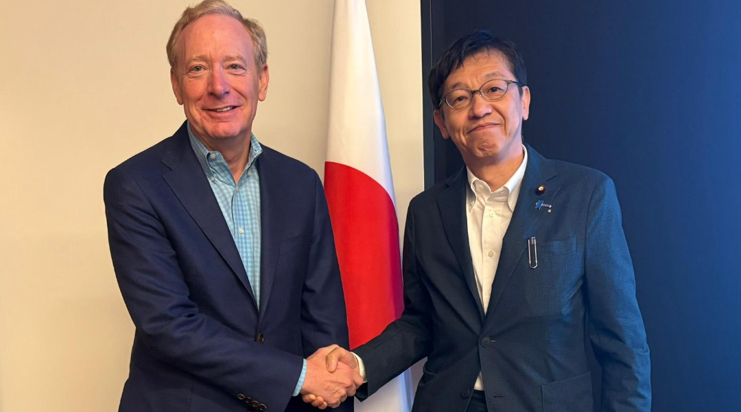 日本の国旗の前で、ブラッド・スミス副会長兼社長と松本デジタル大臣が握手をしている様子。