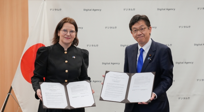 「デジタル庁 / Digital Agency」のロゴが繰り返し印刷された背景に、エストニア共和国のリーサ＝リー・パコスタ法務・デジタル大臣（左）と松本デジタル大臣（右）が署名文書を開いて正面に向け、並んで立っている写真。左側には眼鏡をかけた茶髪のパコスタ法務・デジタル大臣が黒のジャケット（金ボタン）を着用し、胸元にエストニア国旗のピンバッジをつけている。右側には眼鏡をかけた松本デジタル大臣が紺のスーツに青いネクタイを着用し、胸元に日本国旗のピンバッジをつけている。背景左には日本国旗が見える。