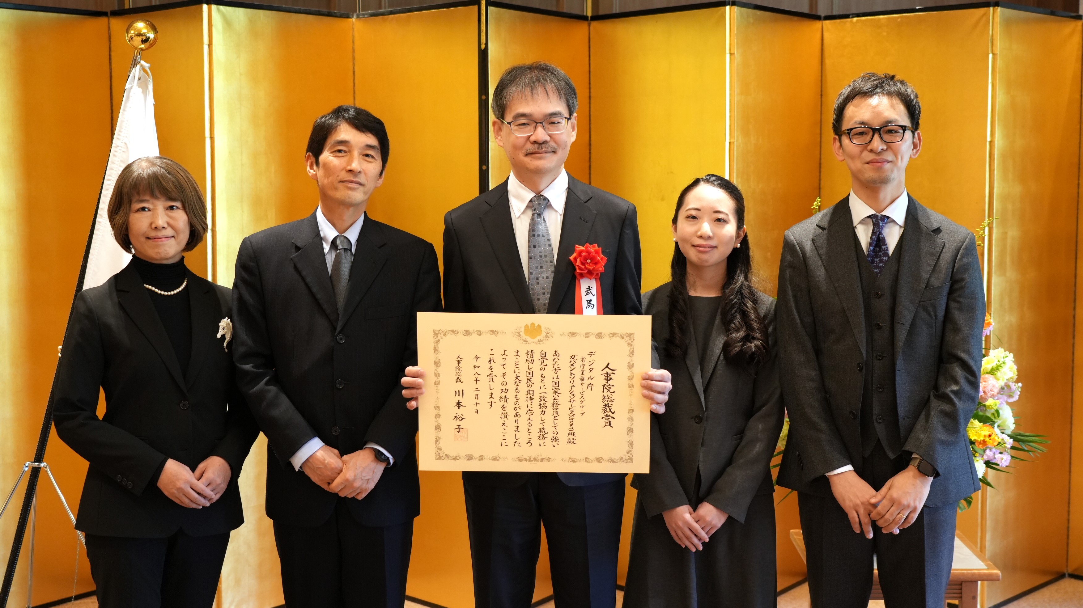 金色の屏風を背景に、三角デジタル監とGSS班職員の計5名がフォーマルな服装で横一列に並び、中央のGSS班職員が表彰状を持って記念撮影をしている
