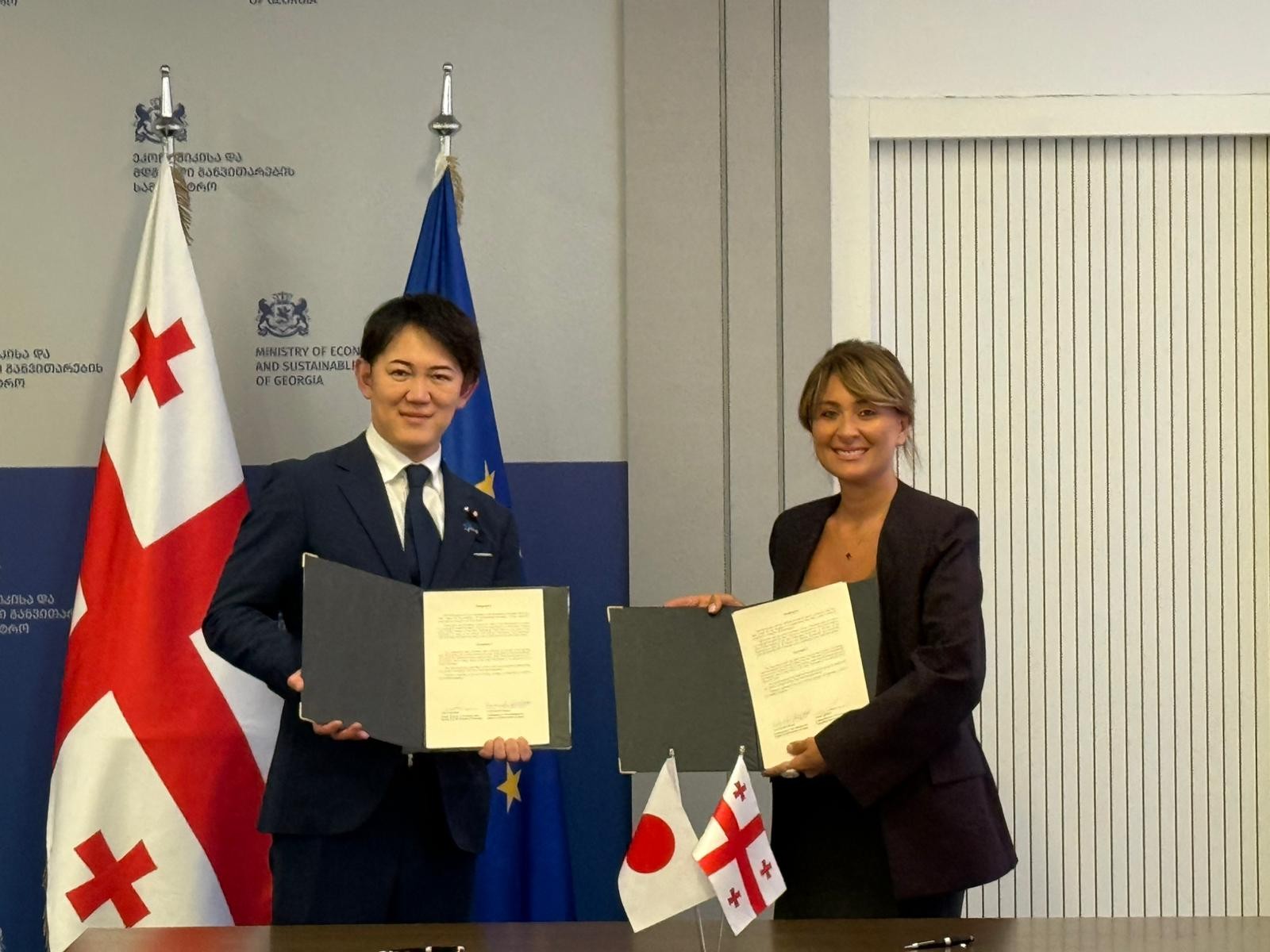 ジョージアと日本の国旗が置かれたデスクの前に協力覚書を持って並んで立つ川崎デジタル政務官とイオセリアニ次官の写真