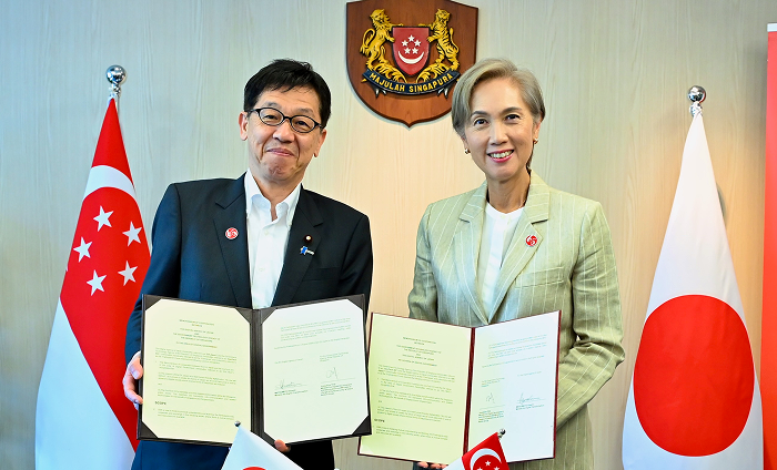 Minister of Digital Transformation Matsumoto and Mrs. Josephine Teo, Minister for Digital Development and Information in Singapore   stand side by side, holding the signed Memorandum of Cooperation. The flags of Japan and Singapore are displayed on either side.