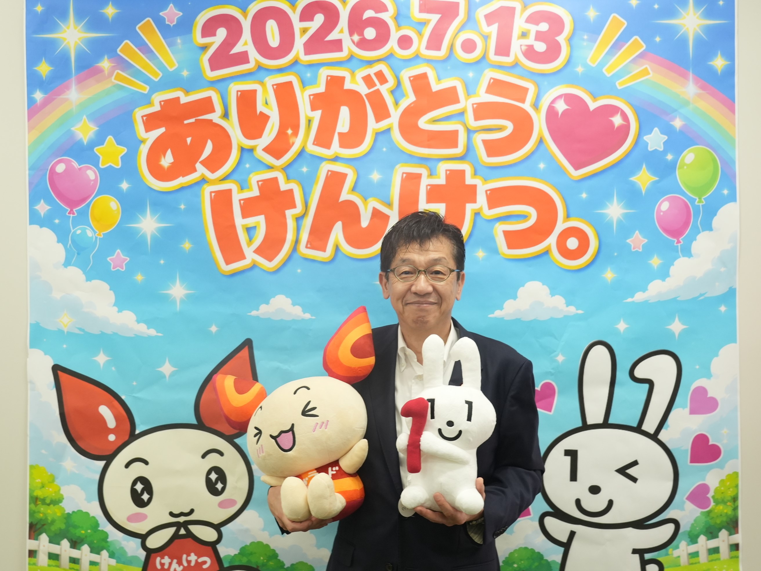 『ありがとう けんけつ。』と書かれたカラフルなポスターの前で、献血キャラクター「けんけつちゃん」とマイナンバーのPRキャラクター「マイナちゃん」のぬいぐるみを両手に抱える松本デジタル大臣